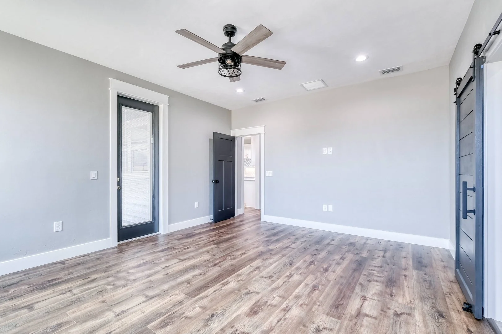 Empty room with light gray walls, wood flooring, a black ceiling fan, a glass door, and a barn door on the right.