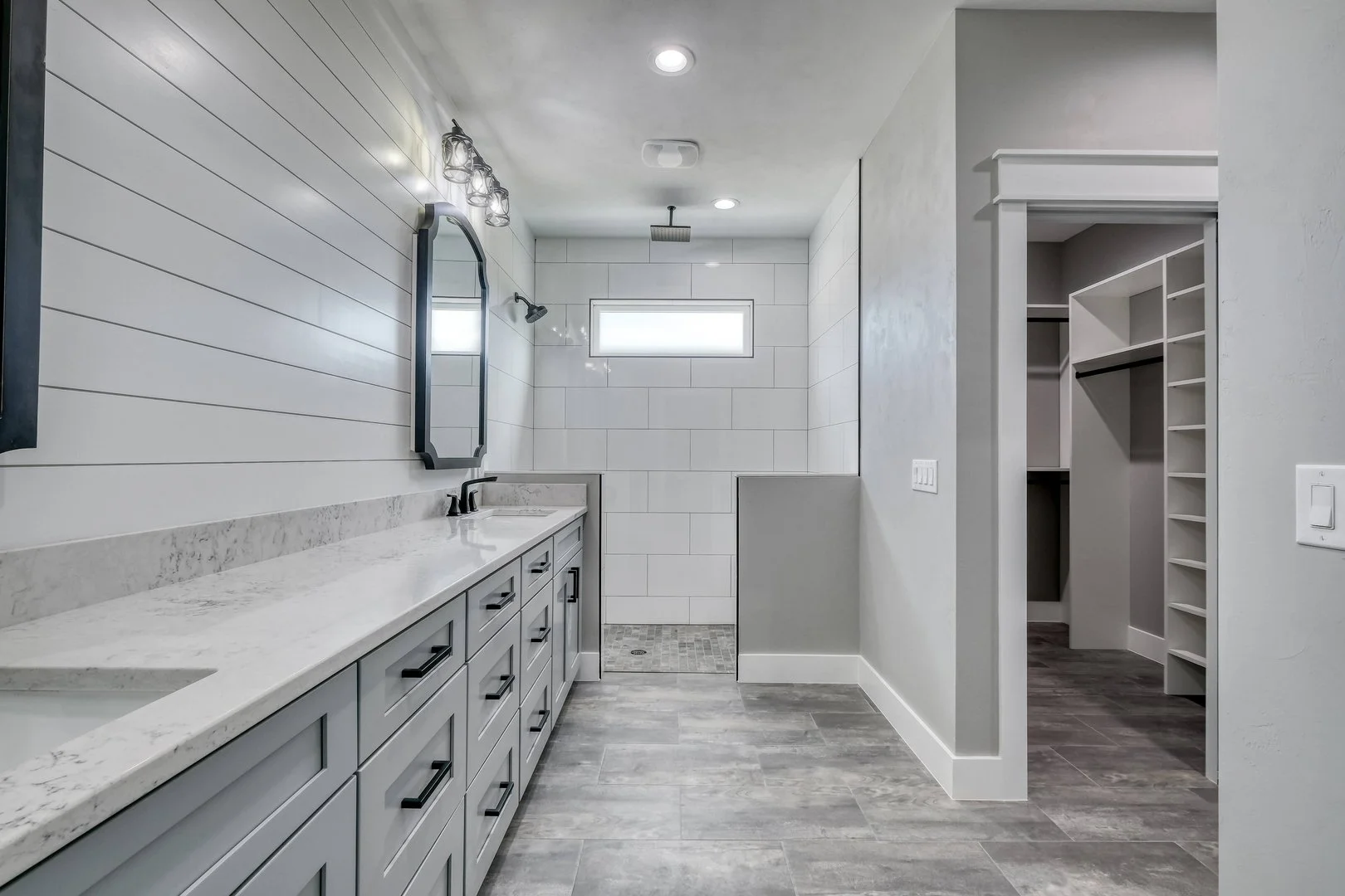 Modern bathroom with gray cabinets, marble countertop, black faucet, mirror, upper vanity lights, white tiled walk-in shower with a horizontal window, and a walk-in closet with white shelving.