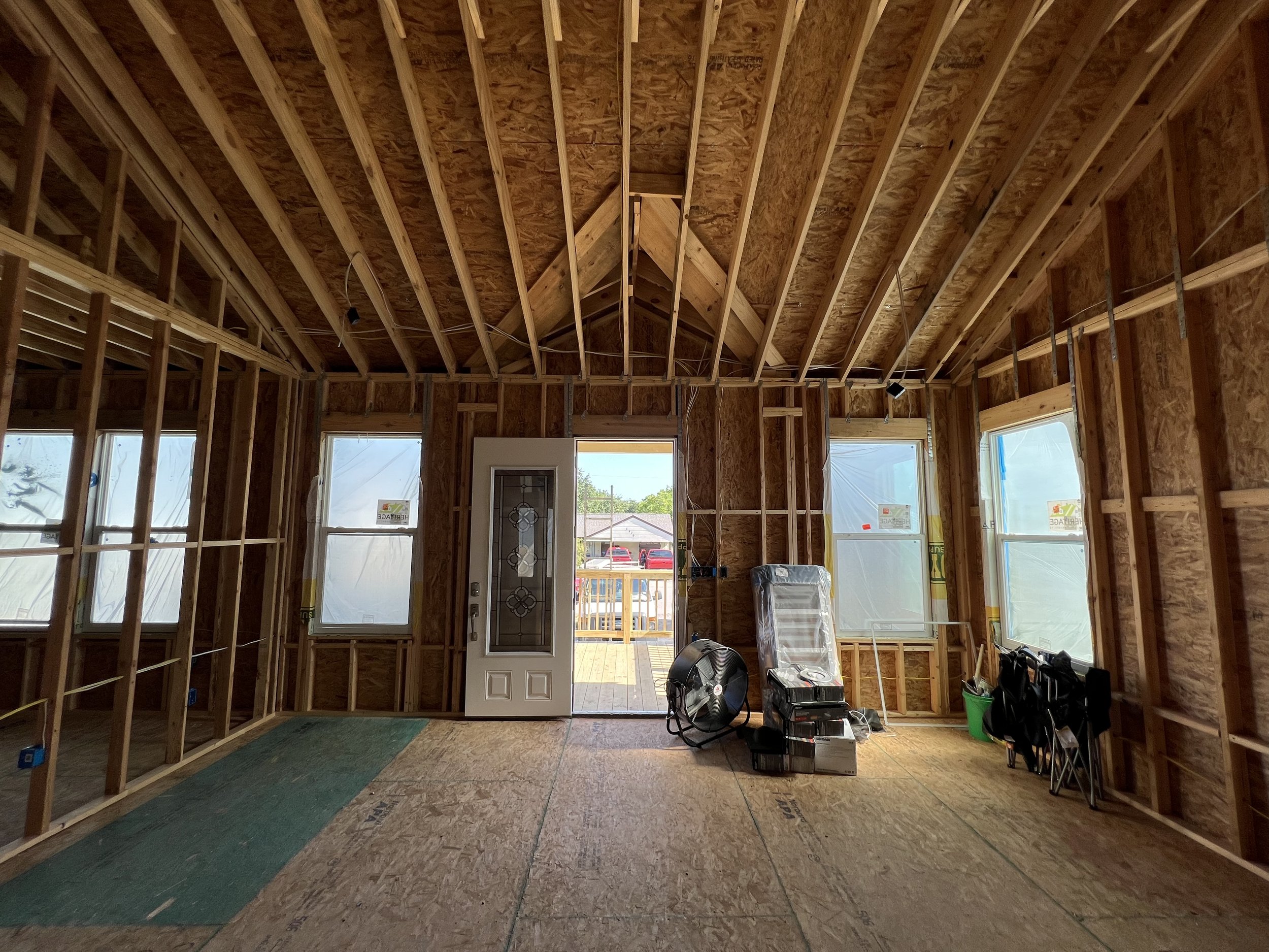 Interior of a house under construction with exposed wooden framing, windows, and a door leading outside. Construction tools and materials are visible on the floor.