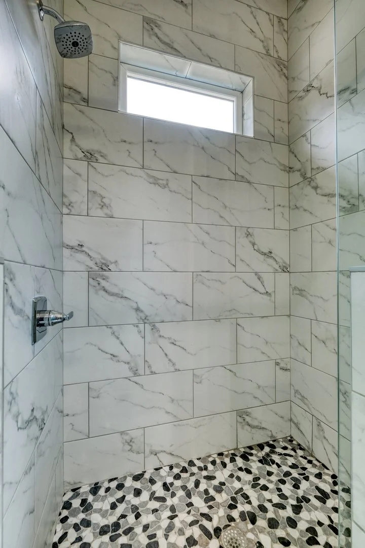 Marble tiled shower with small pebble floor, square window, showerhead, and control knob.