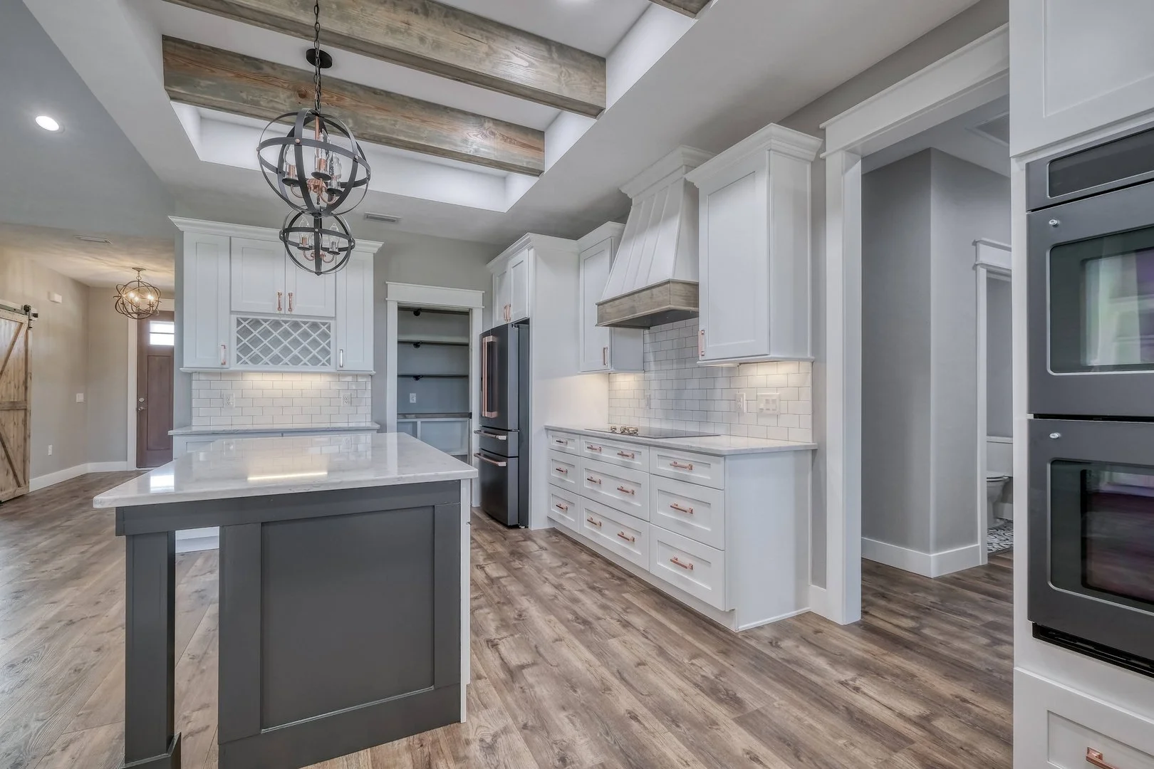 Modern kitchen with white cabinetry, a gray island, wood flooring, and a wood ceiling with exposed beams. The kitchen includes a wine rack, stainless steel appliances, a range hood, and pendant lighting.