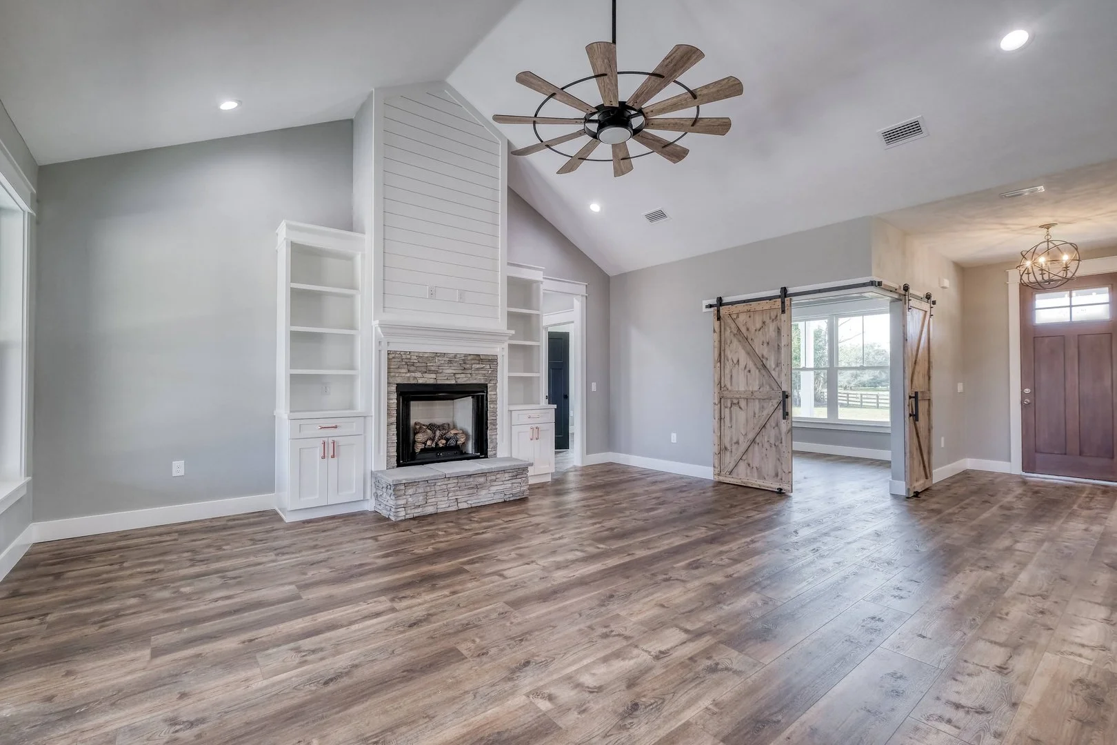 Empty living room with gray walls, wood floor, white built-in shelves around a stone fireplace, ceiling fan, barn door on a sliding track, and a front door with a small window.