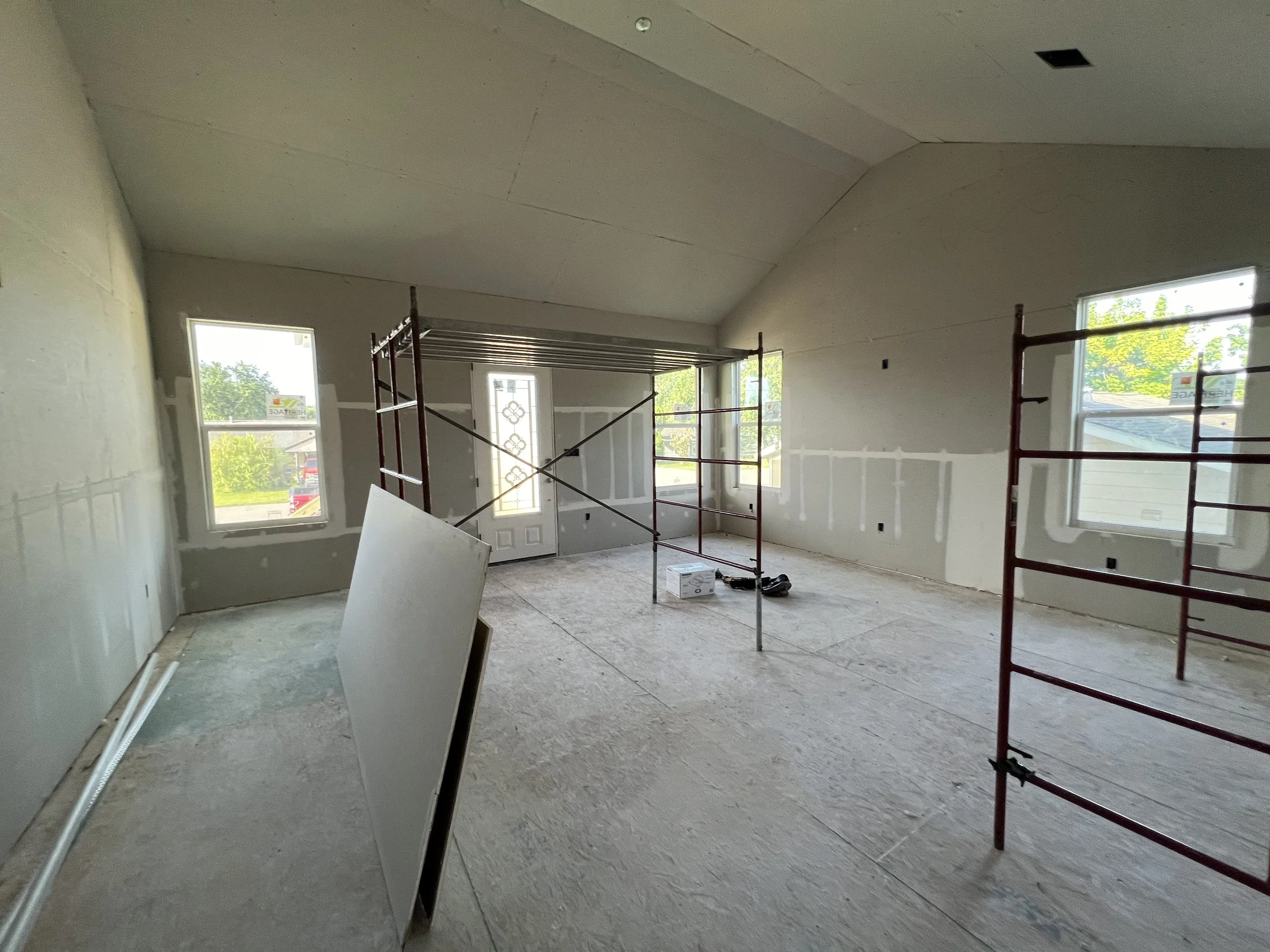 Interior of a house under construction with unfinished walls, large windows, and scaffolding for painting or drywall work.