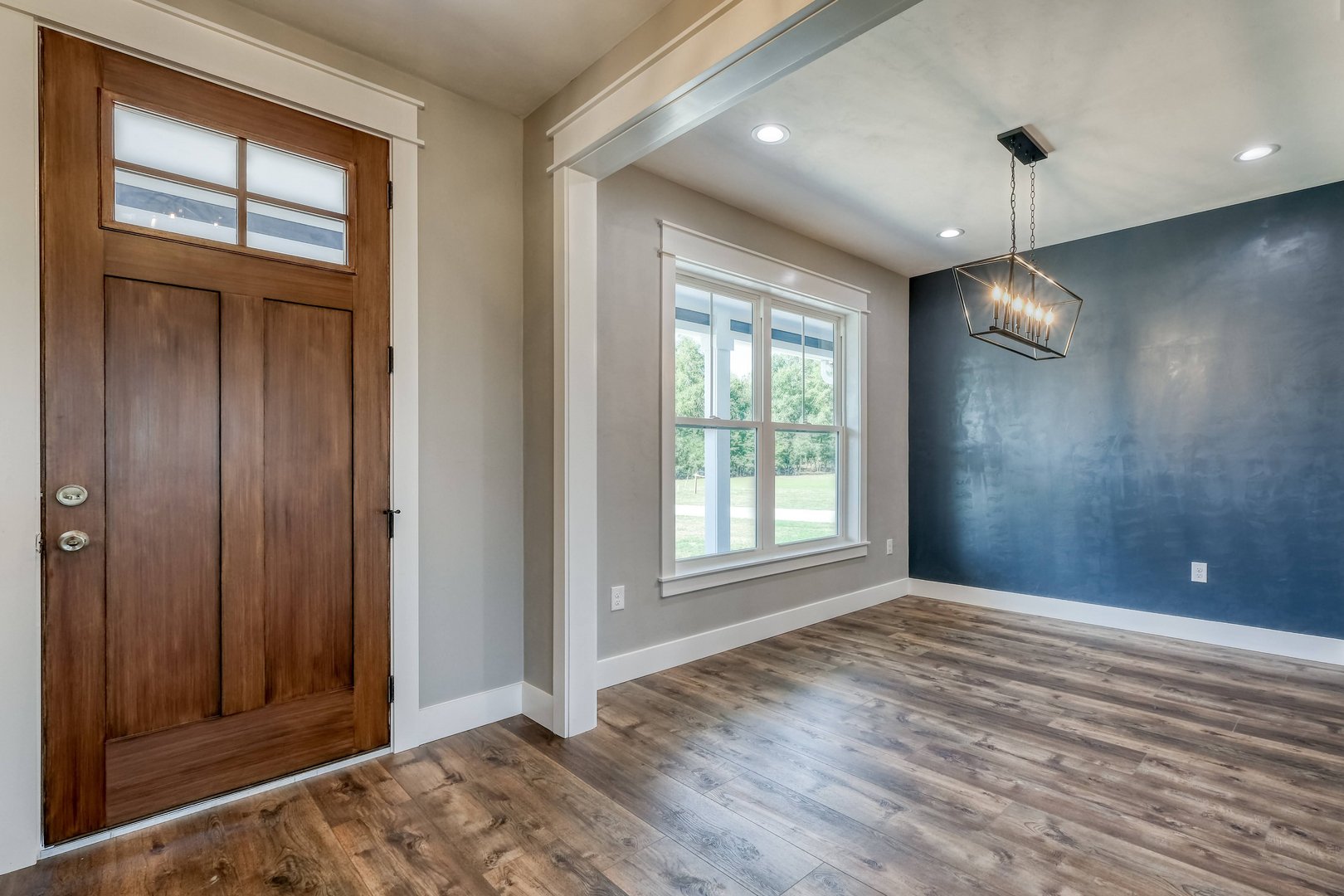 Empty living room with wooden front door, large window, blue accent wall, wood flooring, modern chandelier, and recessed ceiling lights.