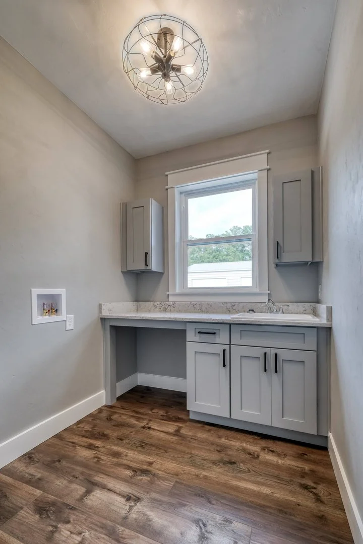 Small laundry room with a window, gray cabinetry, a granite countertop, and a hardwood floor, illuminated by a modern ceiling light fixture.