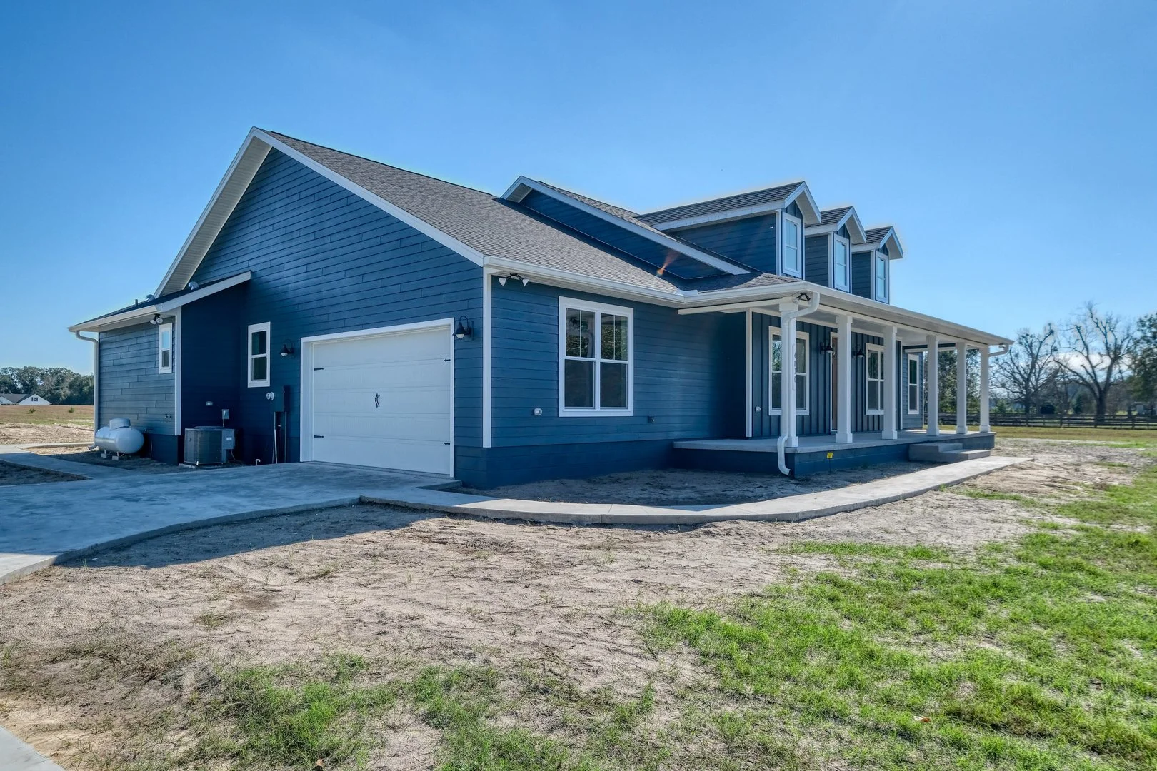 Newly constructed blue house with white trim, attached garage, and front porch on a partly landscaped lot under a clear blue sky.