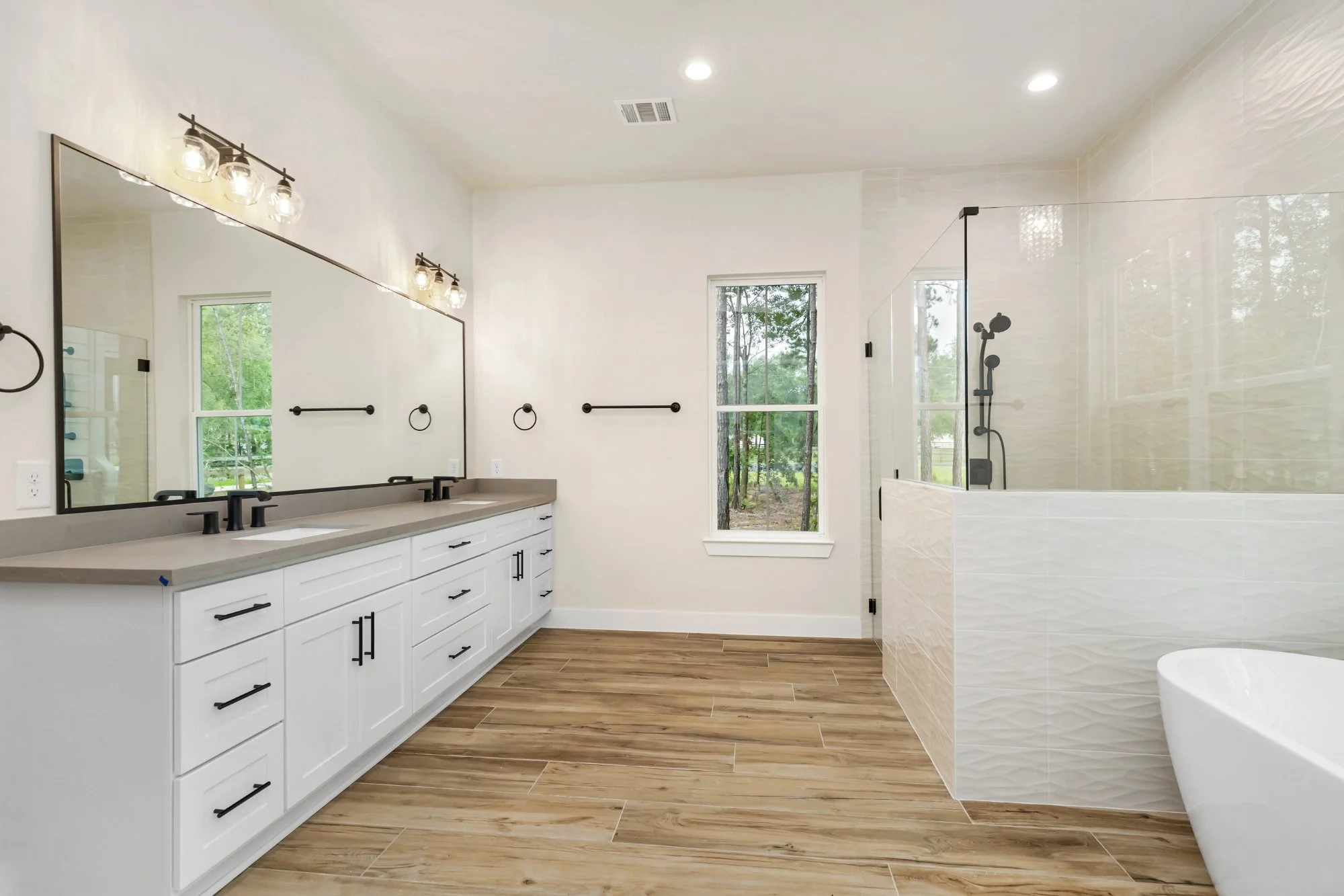Modern bathroom with white vanity, large mirror, black fixtures, a walk-in shower with glass enclosure, and a freestanding bathtub. Wood-look floor tiles and a window providing natural light.