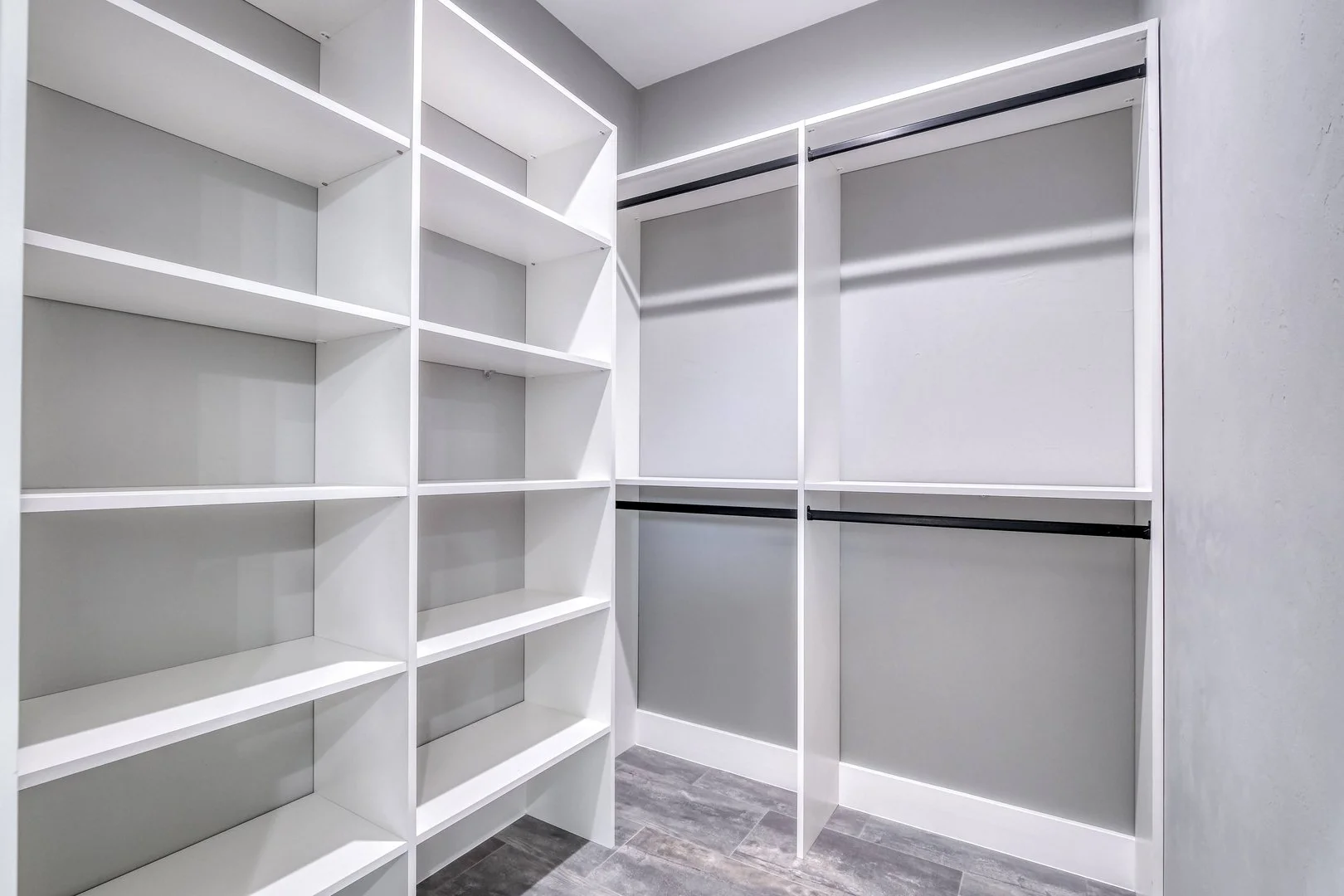 Empty walk-in closet with white shelving units and hanging rods, gray walls, and wood-look flooring.