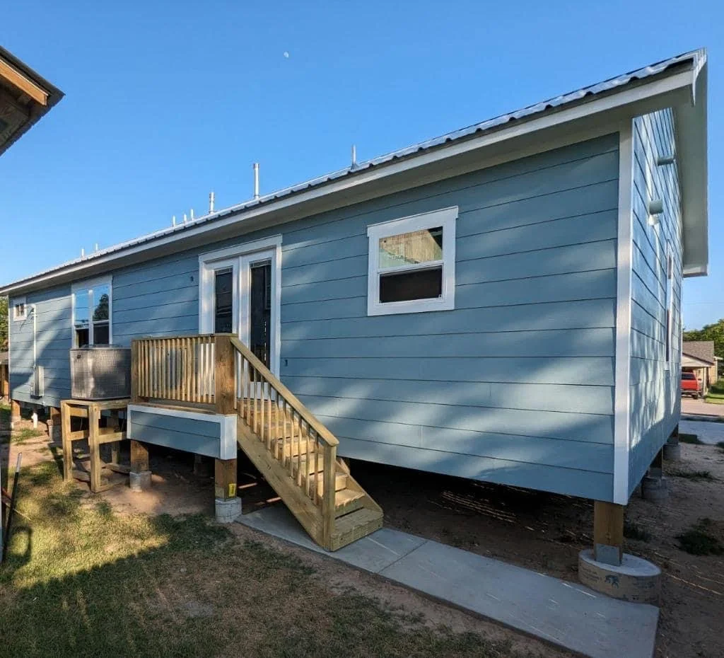 A blue house with a small wooden staircase leading up to a door, and a window on the side. The house is elevated on concrete blocks and has a metal roof.