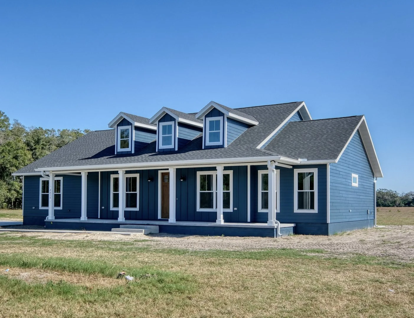 New blue house with white trim, multiple windows, and a front porch, set on a grassy field under a clear blue sky.