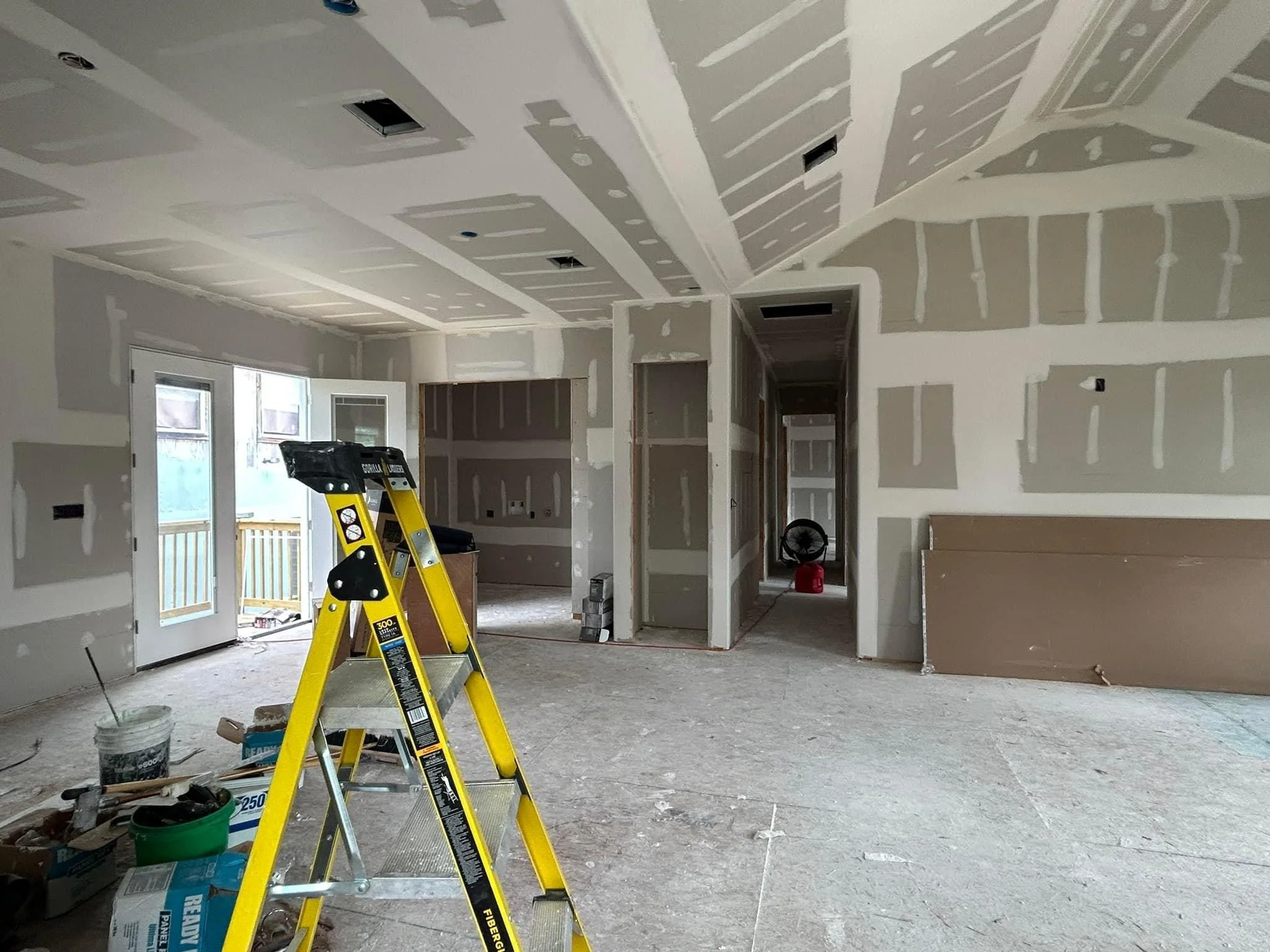 Interior room under construction with drywall installed on walls and ceiling, a yellow ladder, construction materials, and a door leading outside.