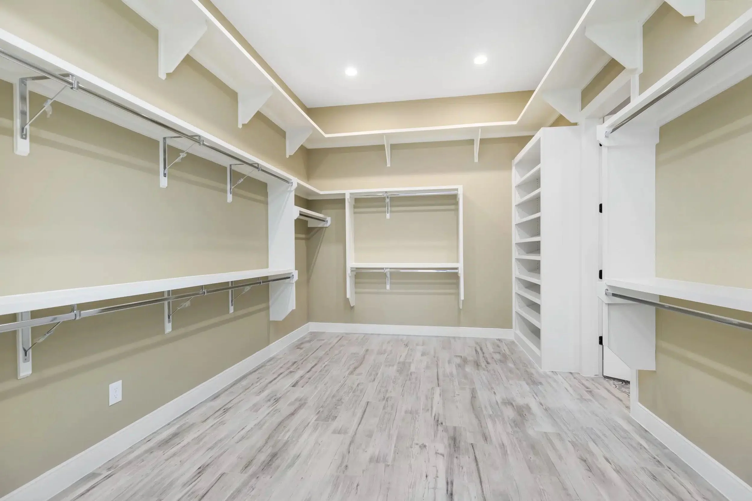 Empty walk-in closet with beige walls, white shelving units, and light wood laminate flooring.