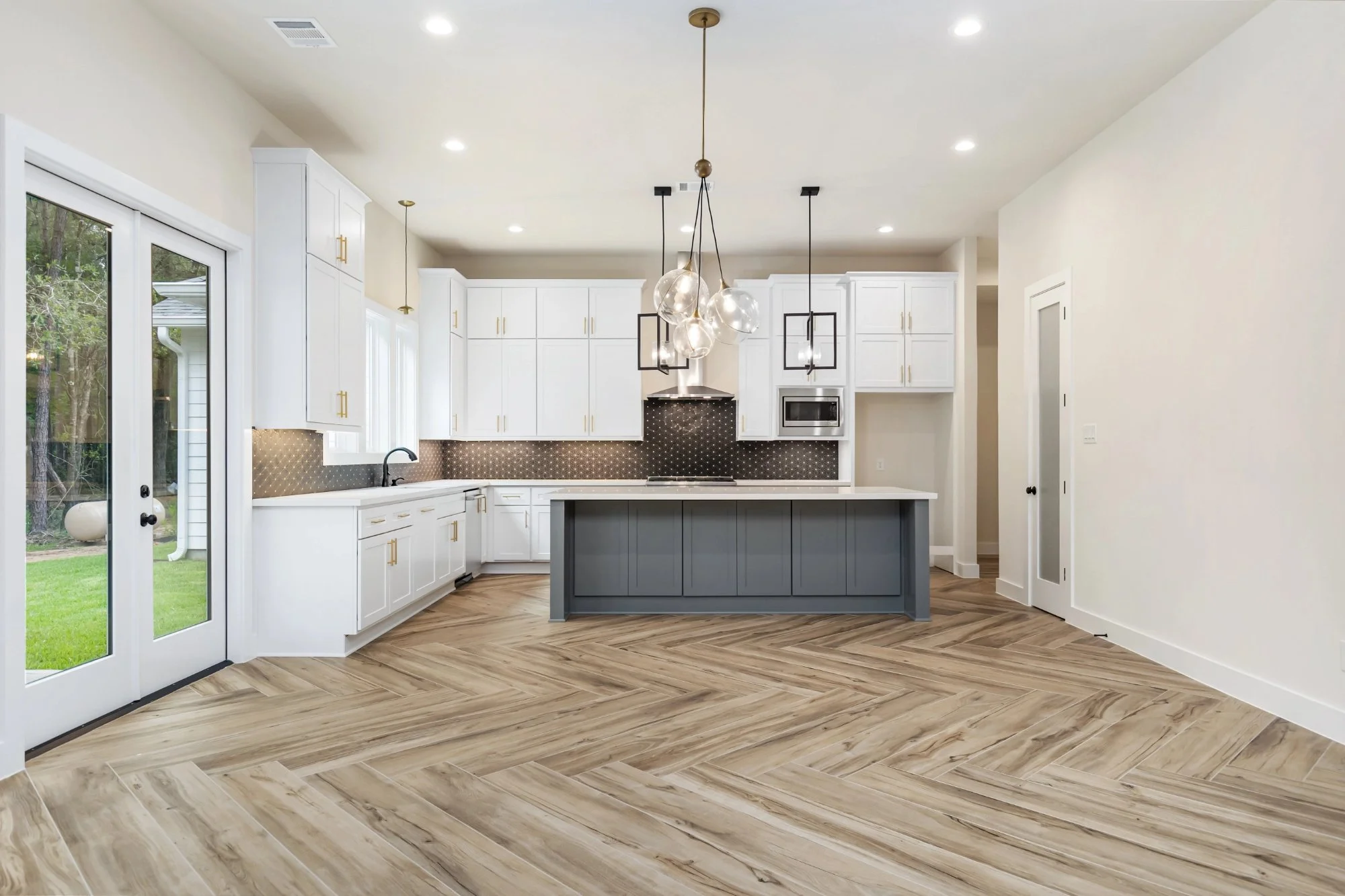 Modern kitchen with white cabinets, black hardware, a gray island with a white countertop, wood-look flooring, and pendant lights with glass globes. There is a slide door leading outside to a backyard.