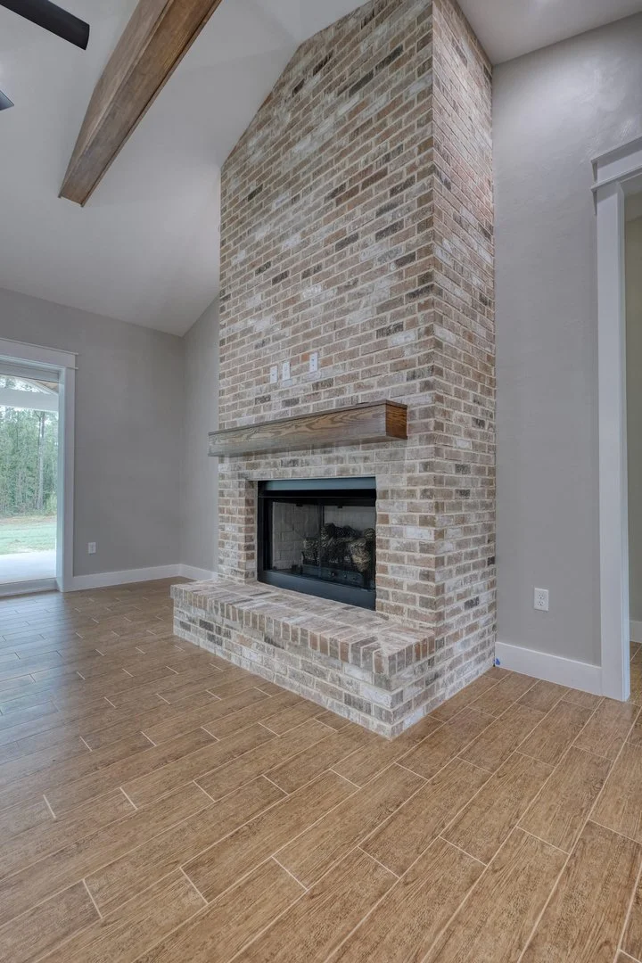 Interior of a modern house with a brick fireplace and wooden mantel, light gray walls, hardwood flooring, and sliding glass door leading to outside greenery.