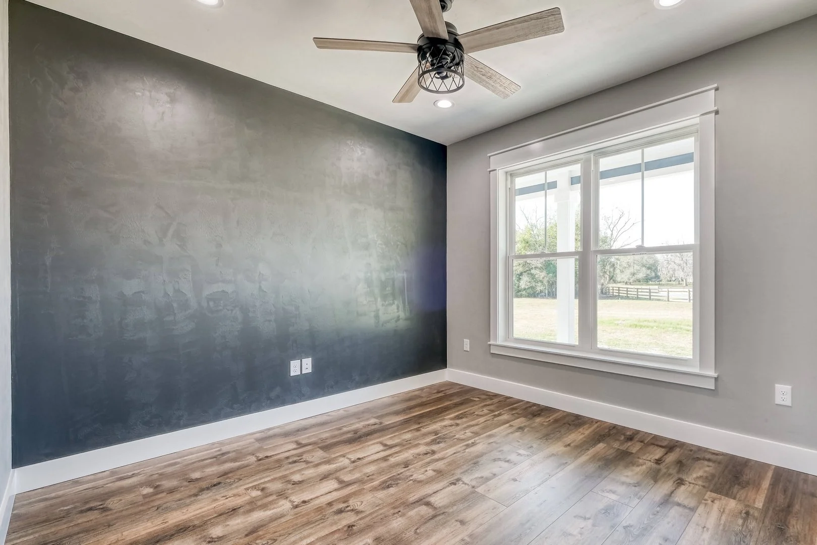 Empty room with a large window, a dark accent wall, wooden flooring, a ceiling fan, and electrical outlets.
