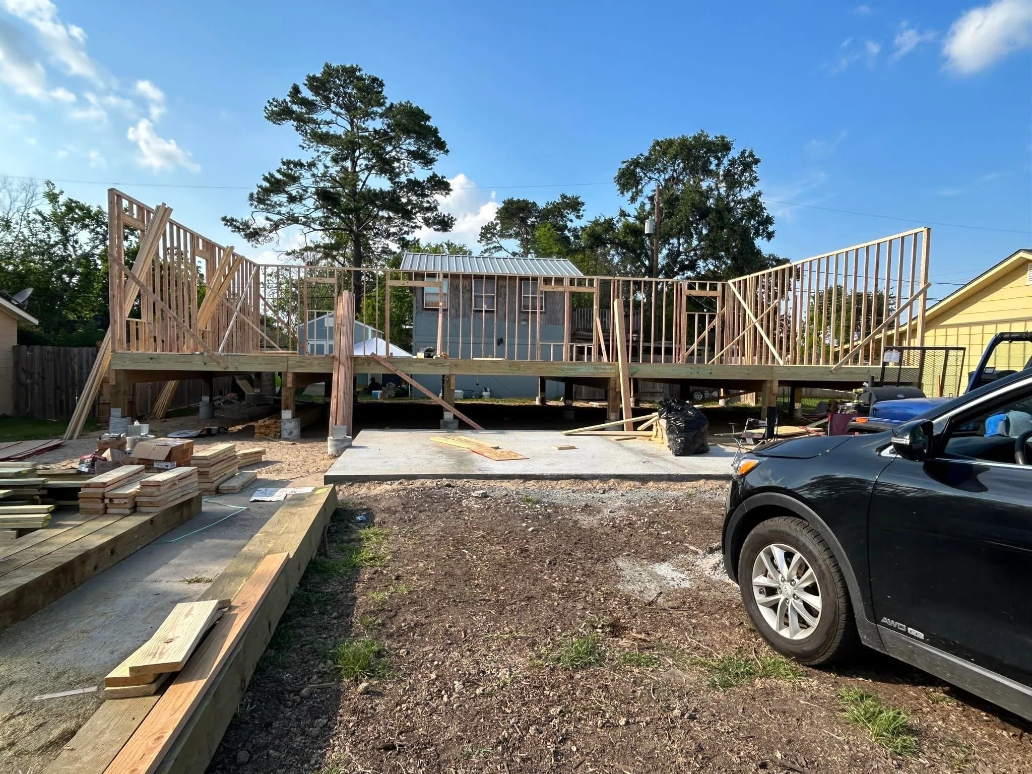 A construction site where a wooden deck or porch is being built, with framing in progress, surrounded by construction materials and tools, and a black car parked nearby.