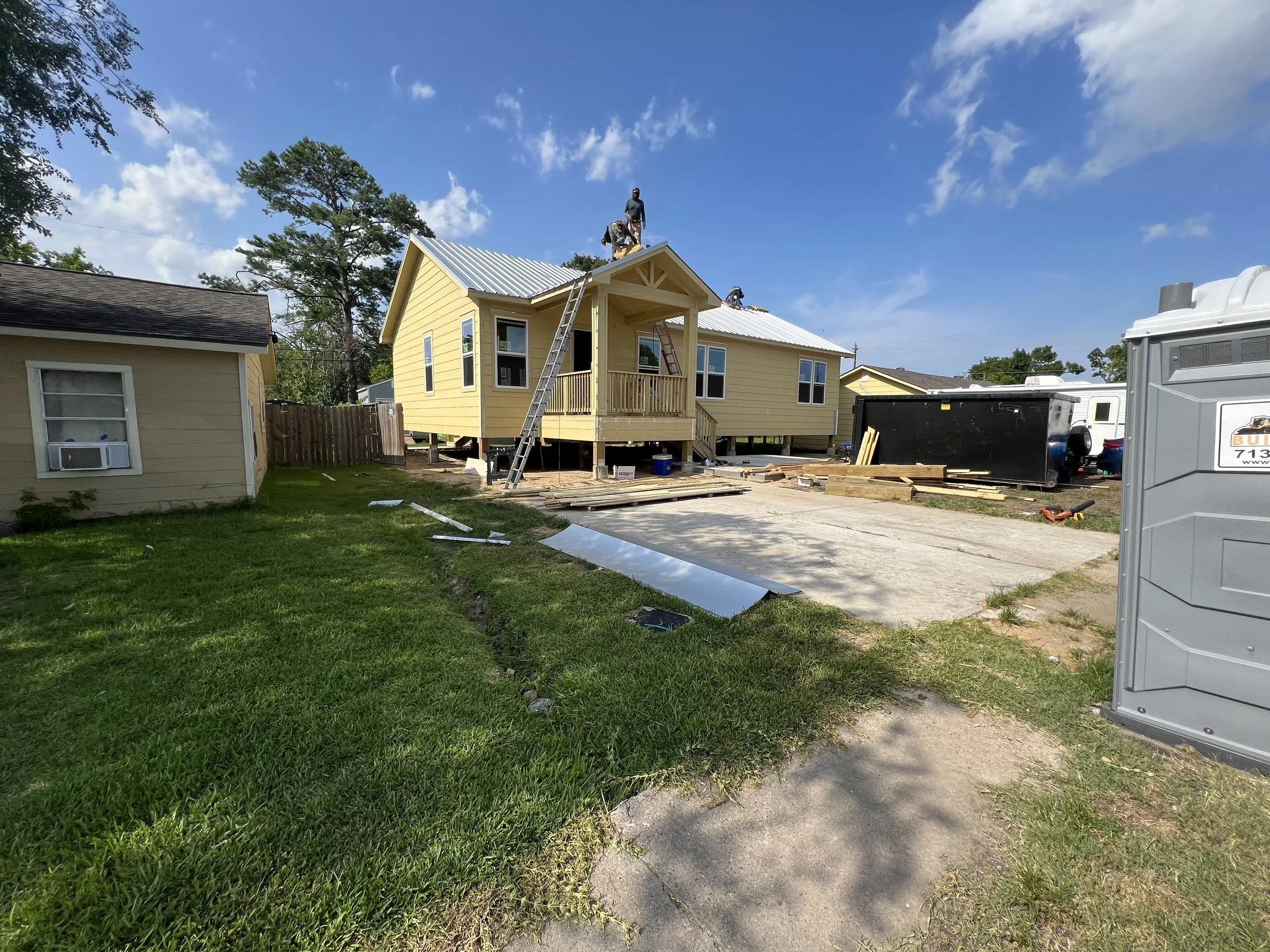 Construction site with workers installing a yellow house on stilts. A ladder leans against the house, with construction materials and tools scattered around. The sky is blue with some clouds.