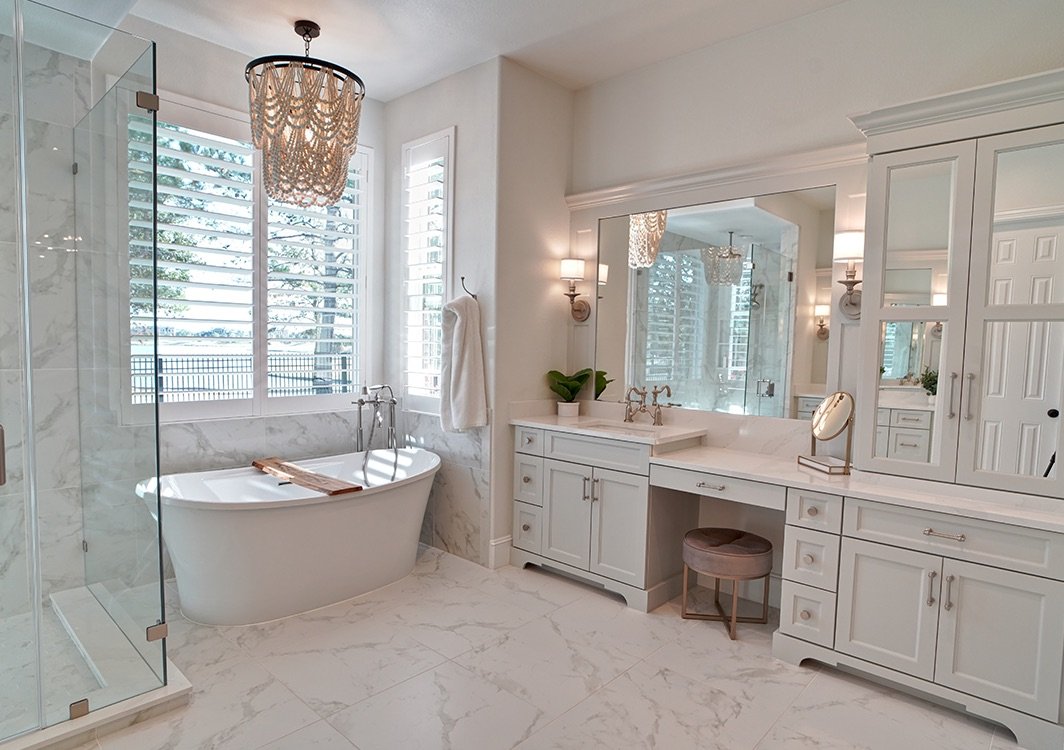 Elegant bathroom with a white freestanding bathtub near large windows with plantation shutters, a glass-enclosed shower, and a white vanity with a large mirror, decorative lighting, and a vanity stool.