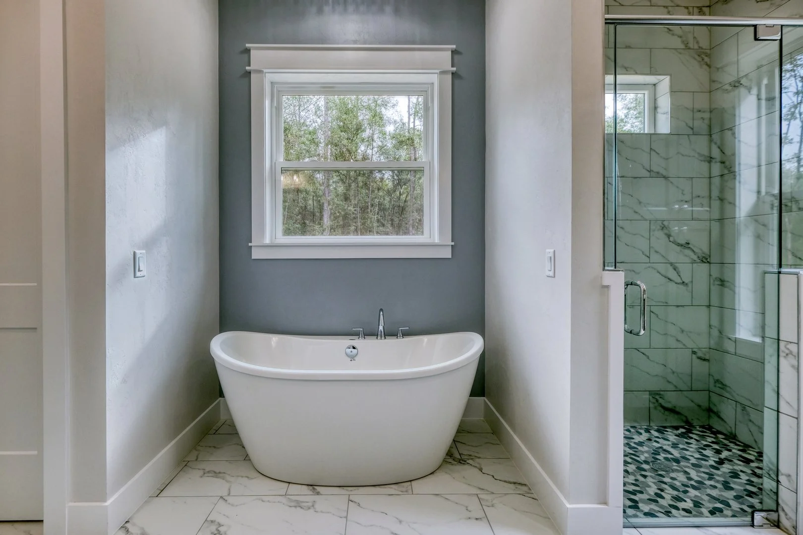Bathroom with a standalone bathtub under a window, adjacent to a walk-in shower with marble tiles and a glass door.