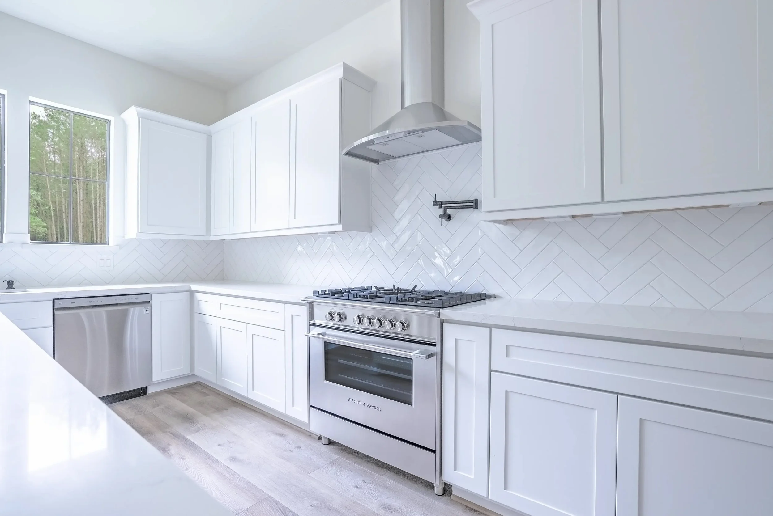 Modern white kitchen with stainless steel appliances, white cabinets, and herringbone tile backsplash, next to a window with a view of trees.