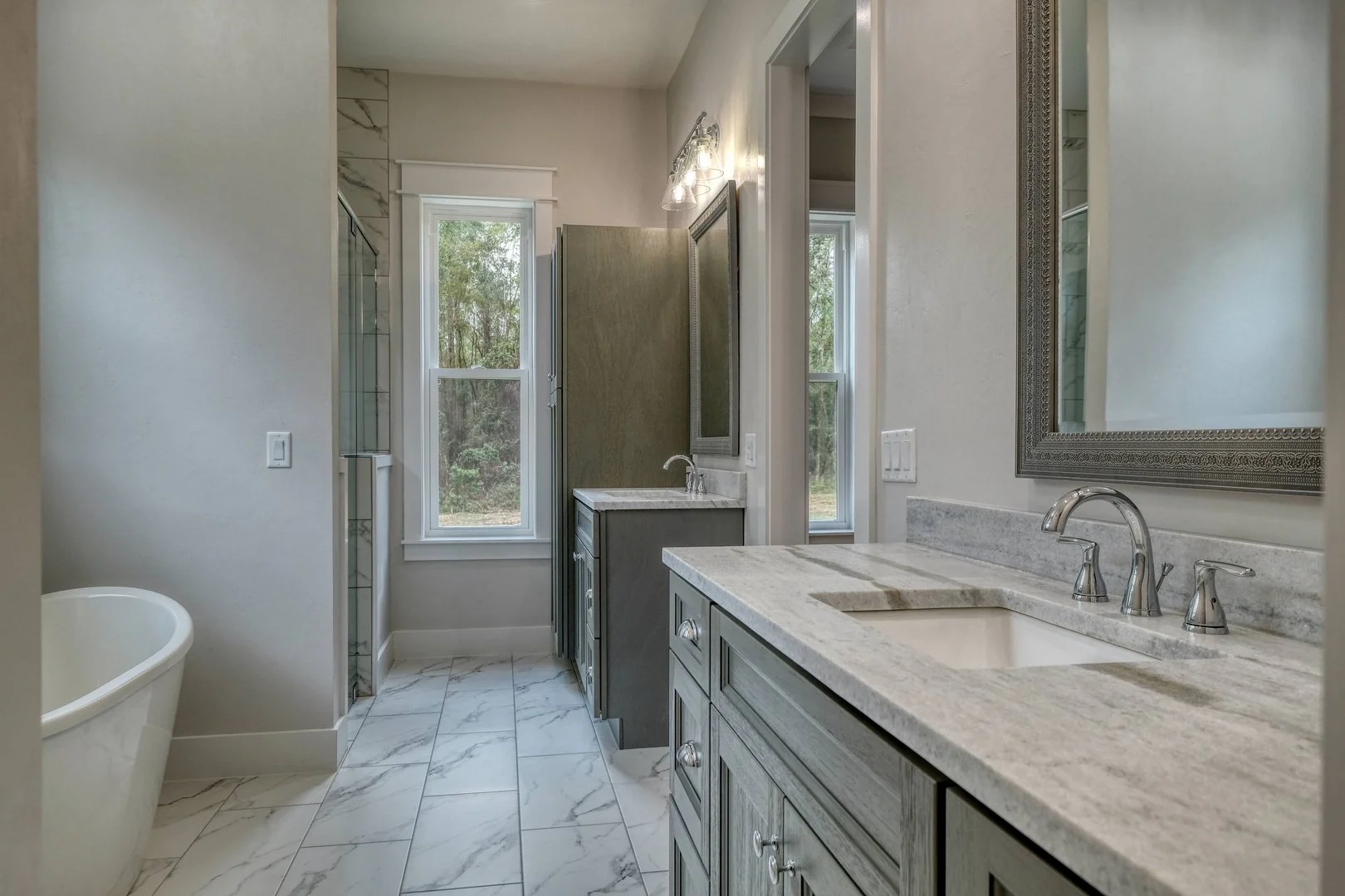 A modern bathroom featuring a gray vanity with a marble countertop, a large mirror, a sink with a chrome faucet, a freestanding bathtub on the left, and a shower with glass doors at the back. There are two windows allowing natural light to illuminate