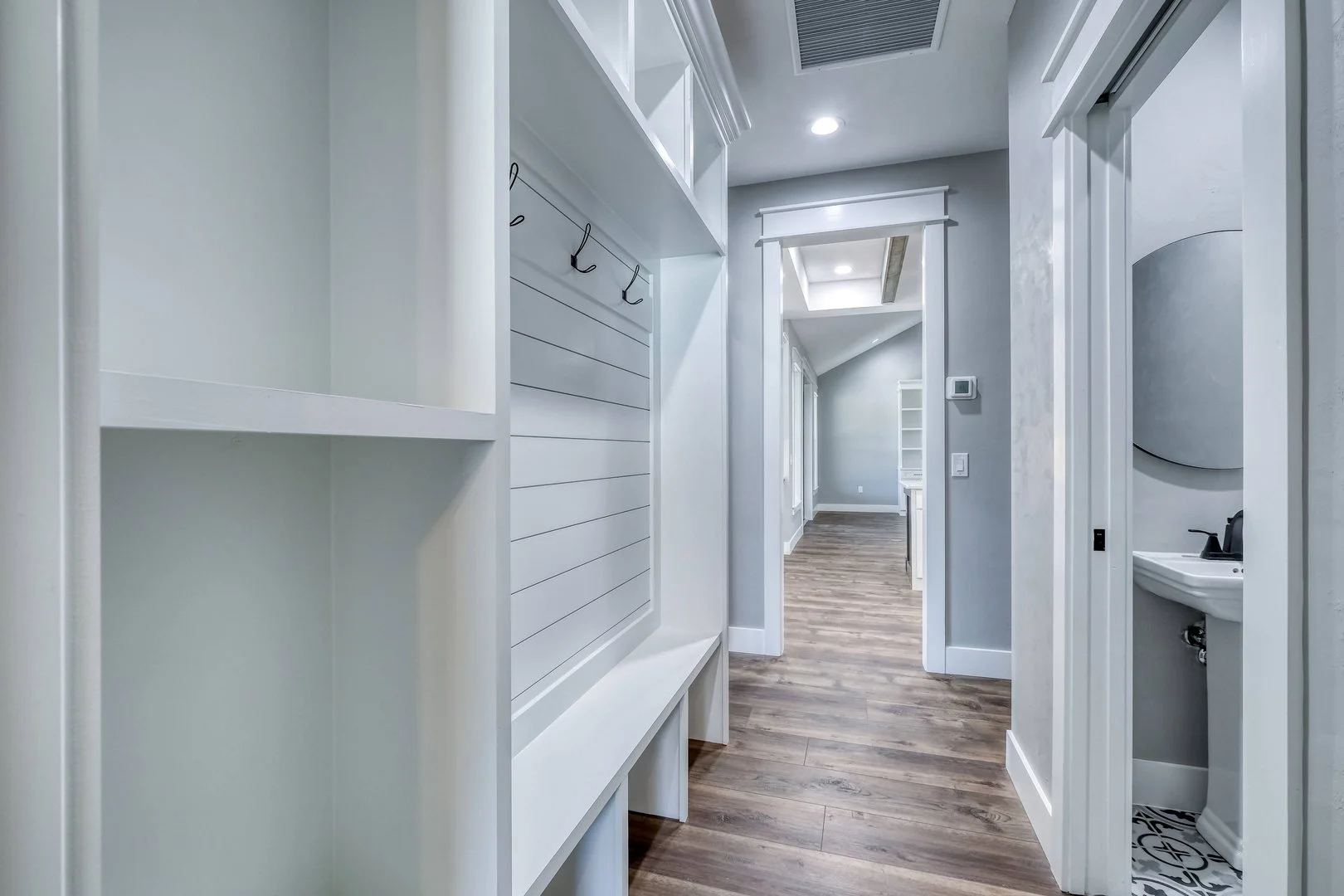 Interior of a modern home hallway with white walls, wood flooring, and built-in shelves and hooks, leading to other rooms and a small bathroom.