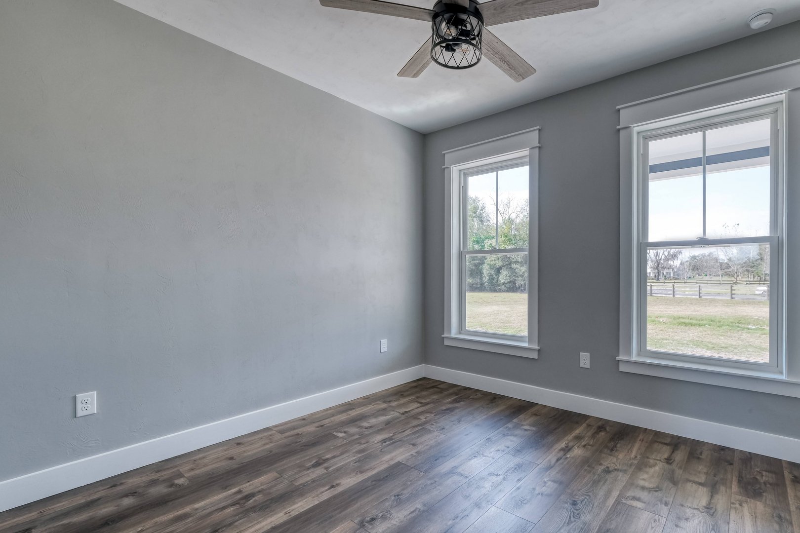 Empty room with gray walls, wooden flooring, two large windows showing an outdoor landscape, and a ceiling fan.