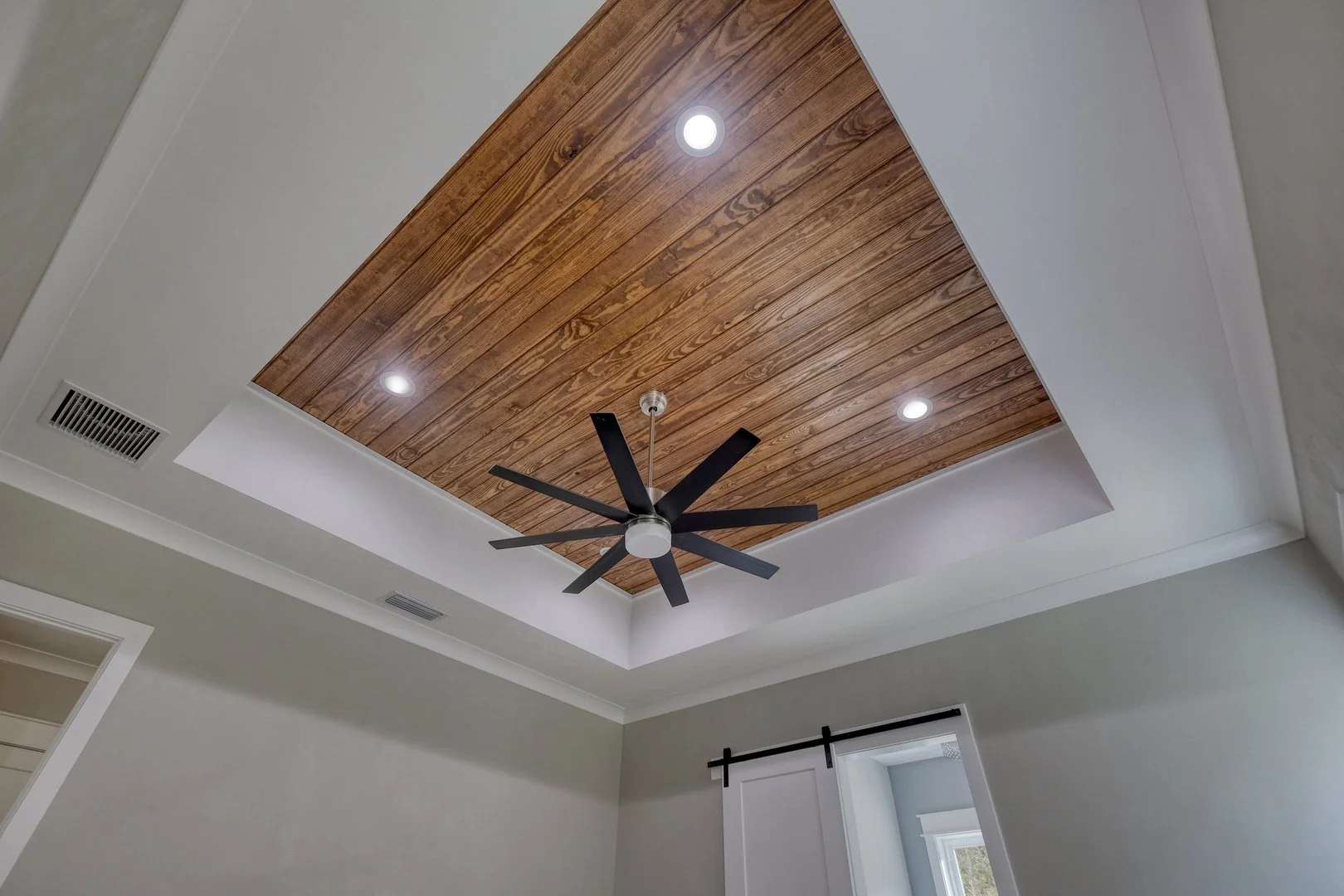 Ceiling with recessed lighting, a wooden panel inset, a ceiling fan, and part of a room with curtains and a window.