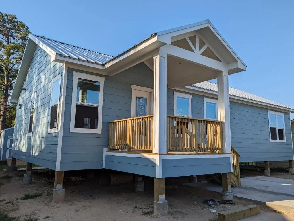A newly built, light blue house with white trim on stilts, featuring a small front porch with a wooden railing, four windows, and a metal roof, under a clear blue sky.