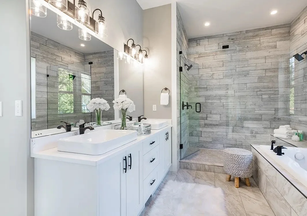 Modern bathroom with double vanity, large mirror, glass-enclosed shower, and bathtub. Decor includes white flowers, towels, and a cushioned stool.