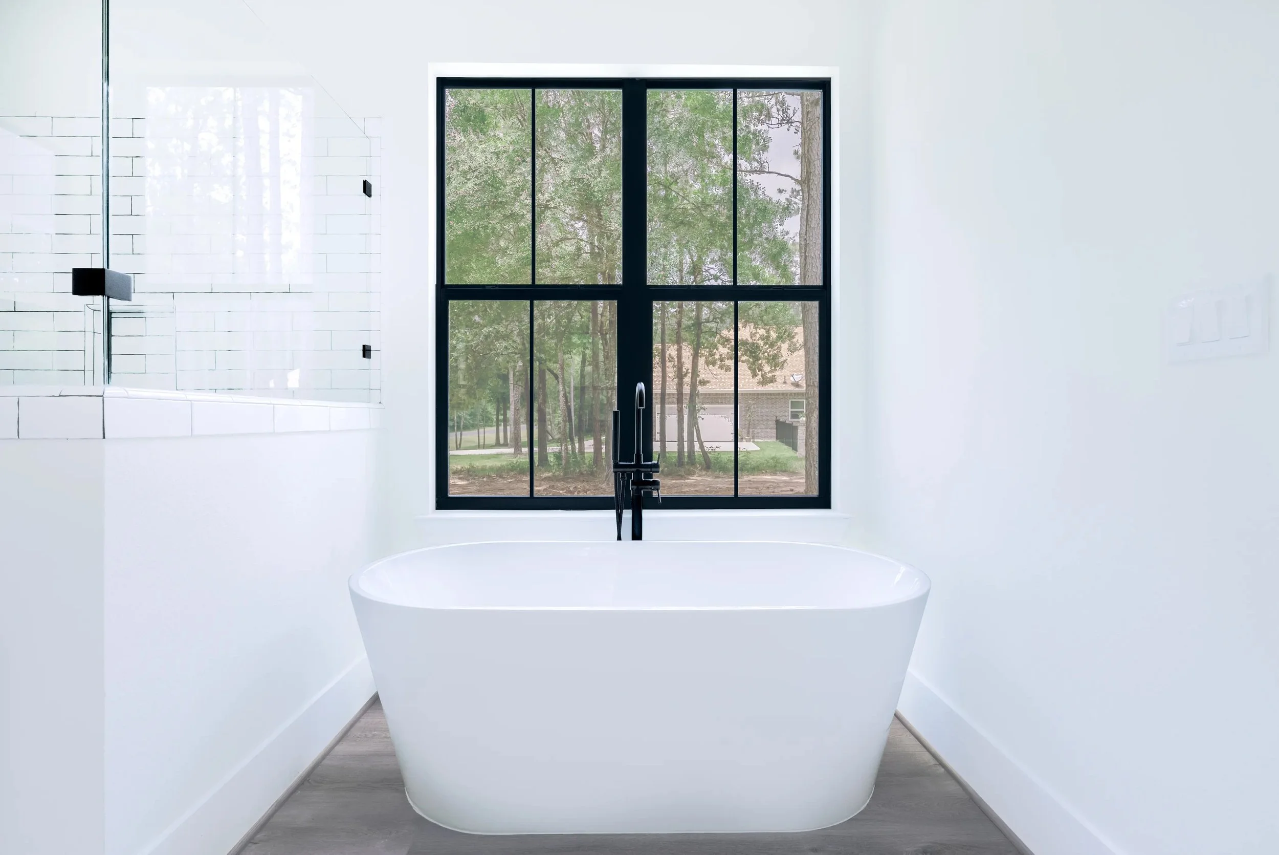 Empty white bathtub in a bright bathroom with large window showing green trees outside.