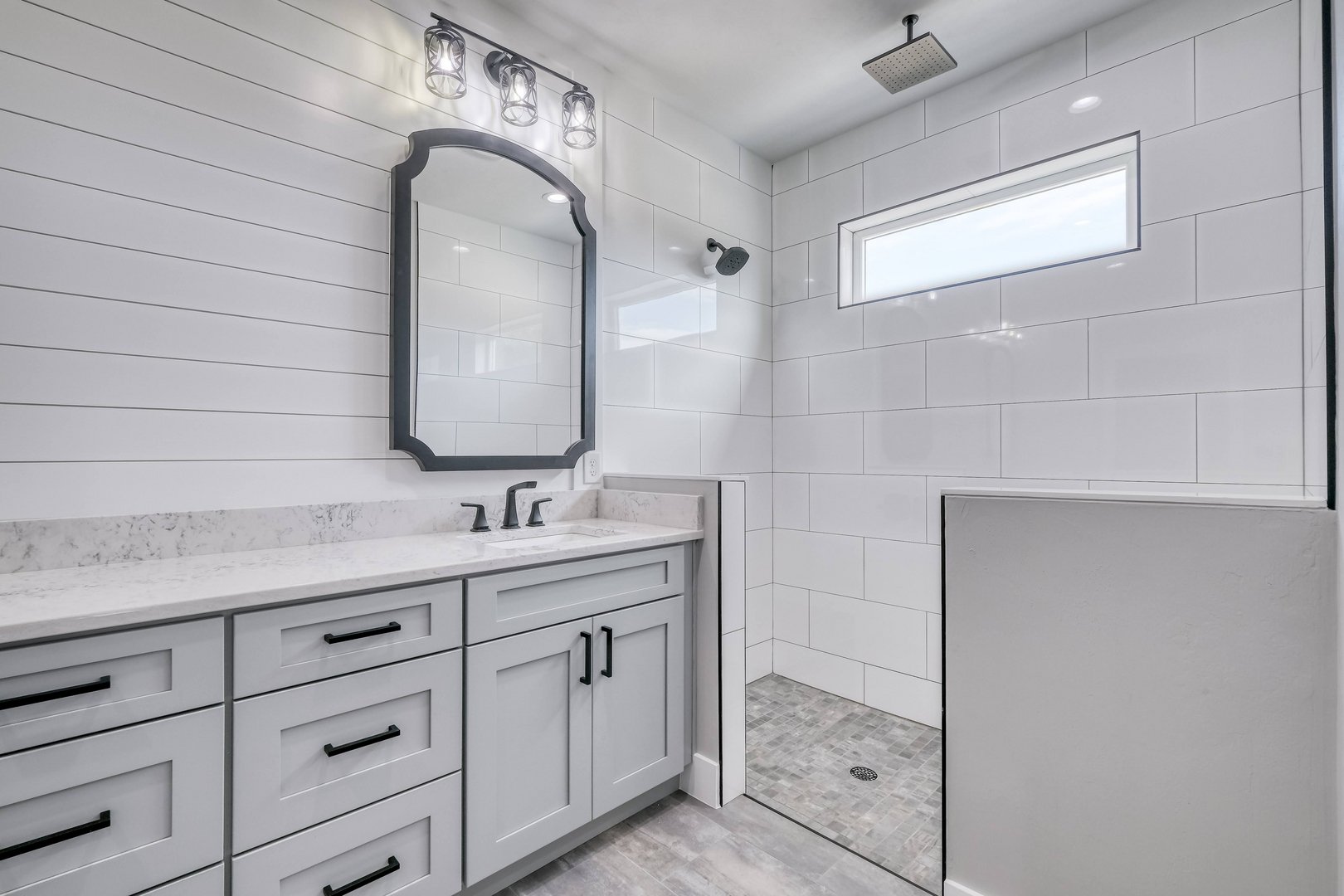 Modern bathroom with white cabinetry, a marble countertop, and a mirror. Features a walk-in shower with white tiles and a small window.