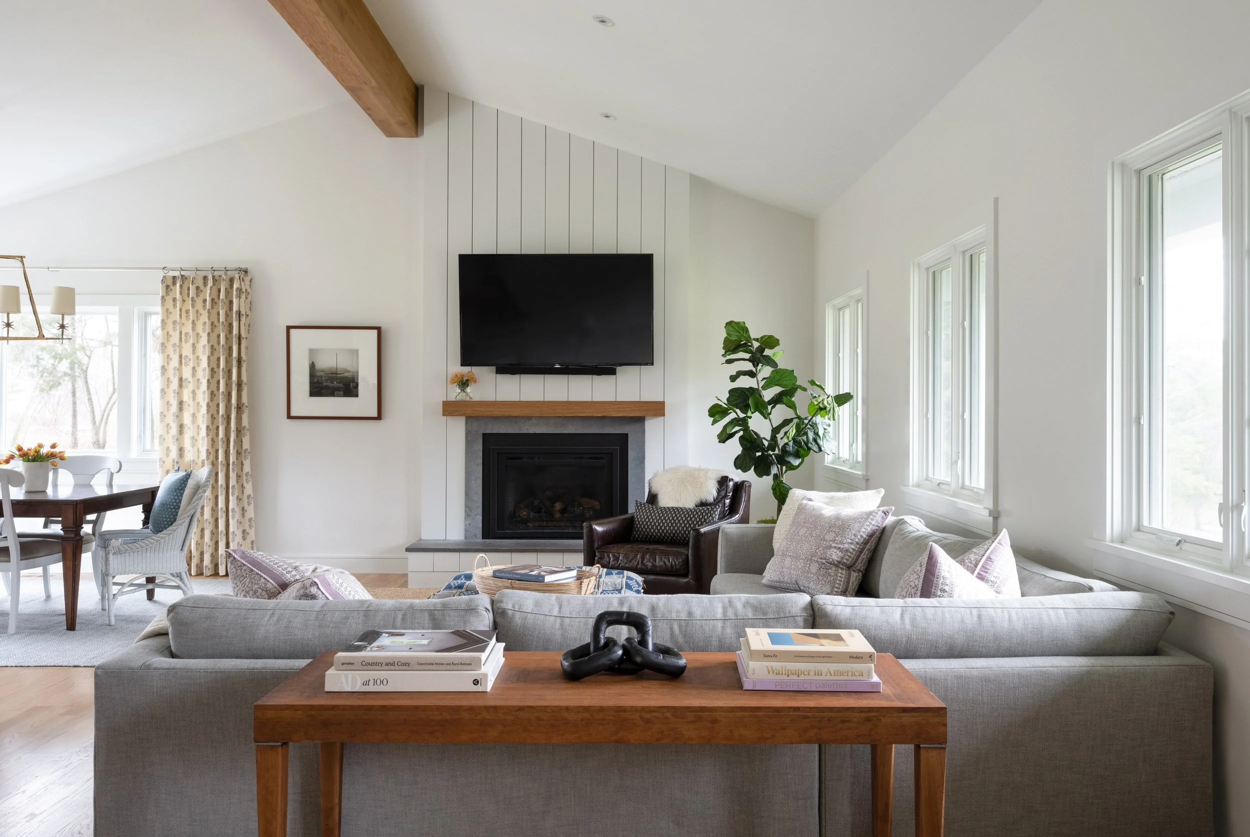 Bright living room with white walls, multiple rectangular windows, a gray sectional sofa with pillows, a wooden coffee table with books and decorative objects, a black leather chair, a fireplace with a wooden mantel, a large flat-screen TV above the fireplace, and a tall green plant.