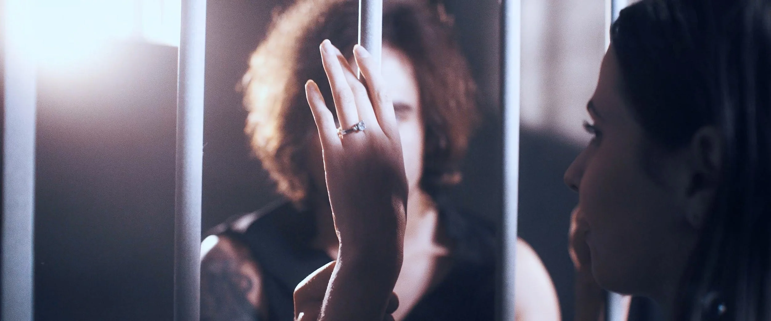 A woman with curly hair touching the glass of a jail cell, with her face close to the glass and her finger pressed against it, while another woman with straight hair looks at her.