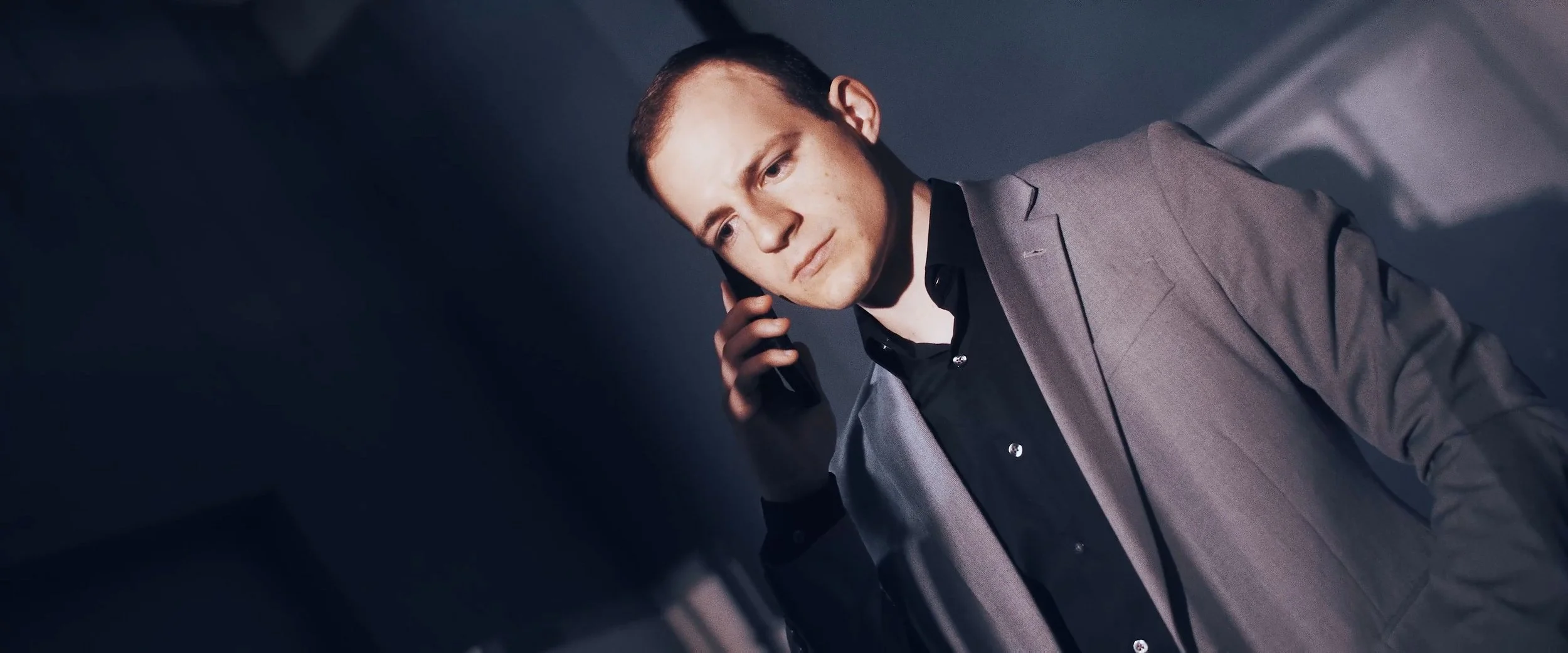 A man in a gray suit and black shirt talking on a cell phone.