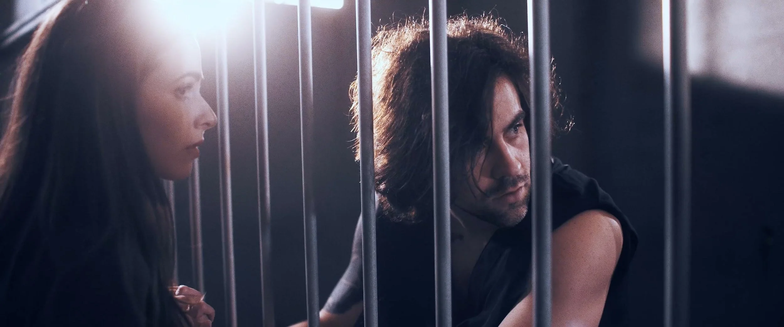A man and woman behind bars, appearing serious or contemplative, in a dimly lit setting.