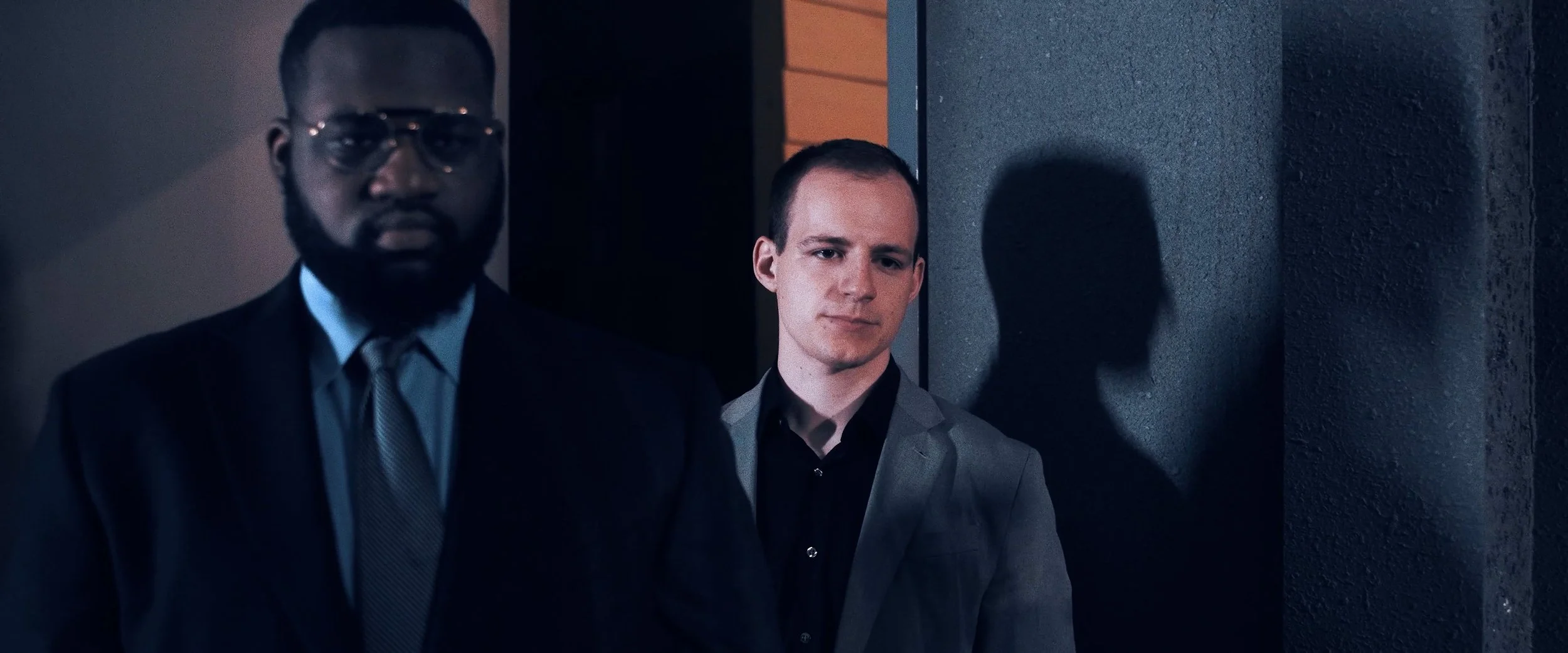 Two men in suits standing near a wall with shadows cast behind them. One man is in front, wearing glasses and a suit with a tie, while the other man is in the background, looking at the camera with a slight smile.