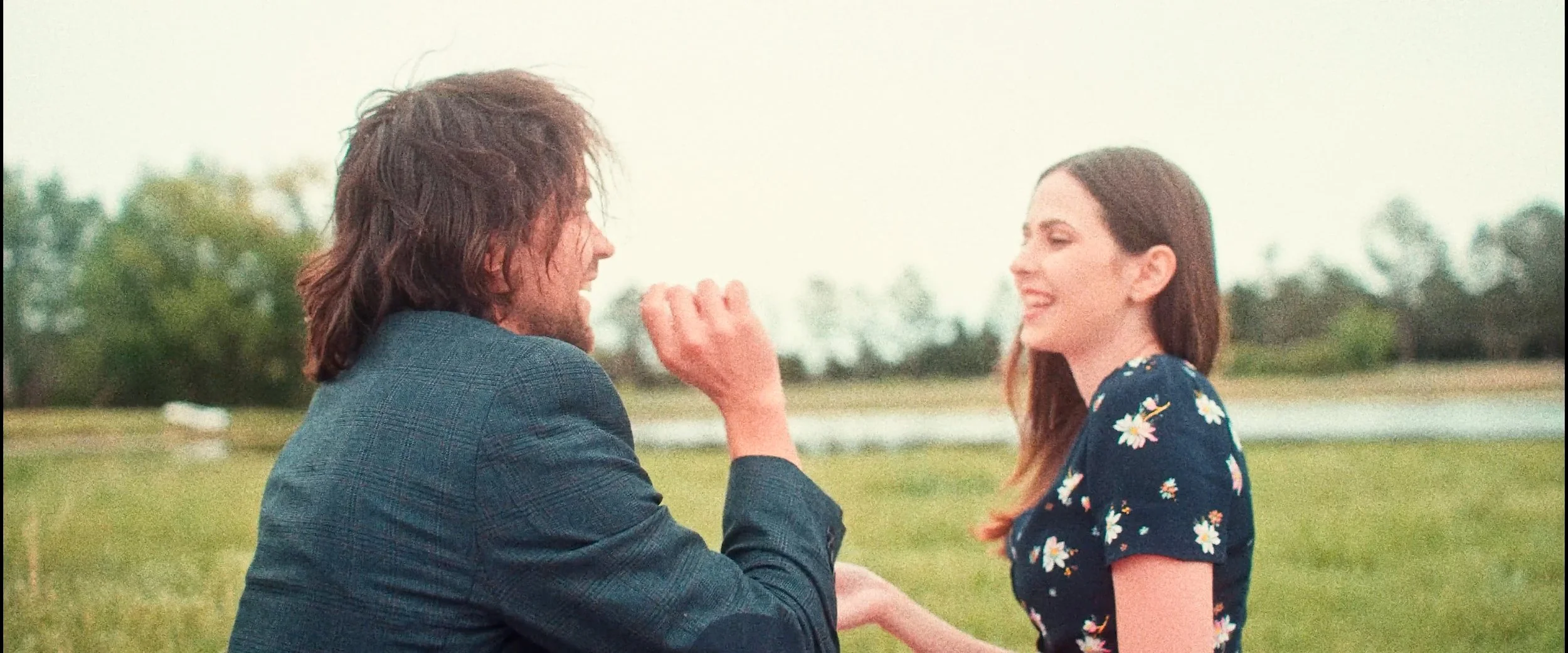 A man kneeling on the grass proposes to a woman, who is smiling, outdoors near a pond with trees in the background.