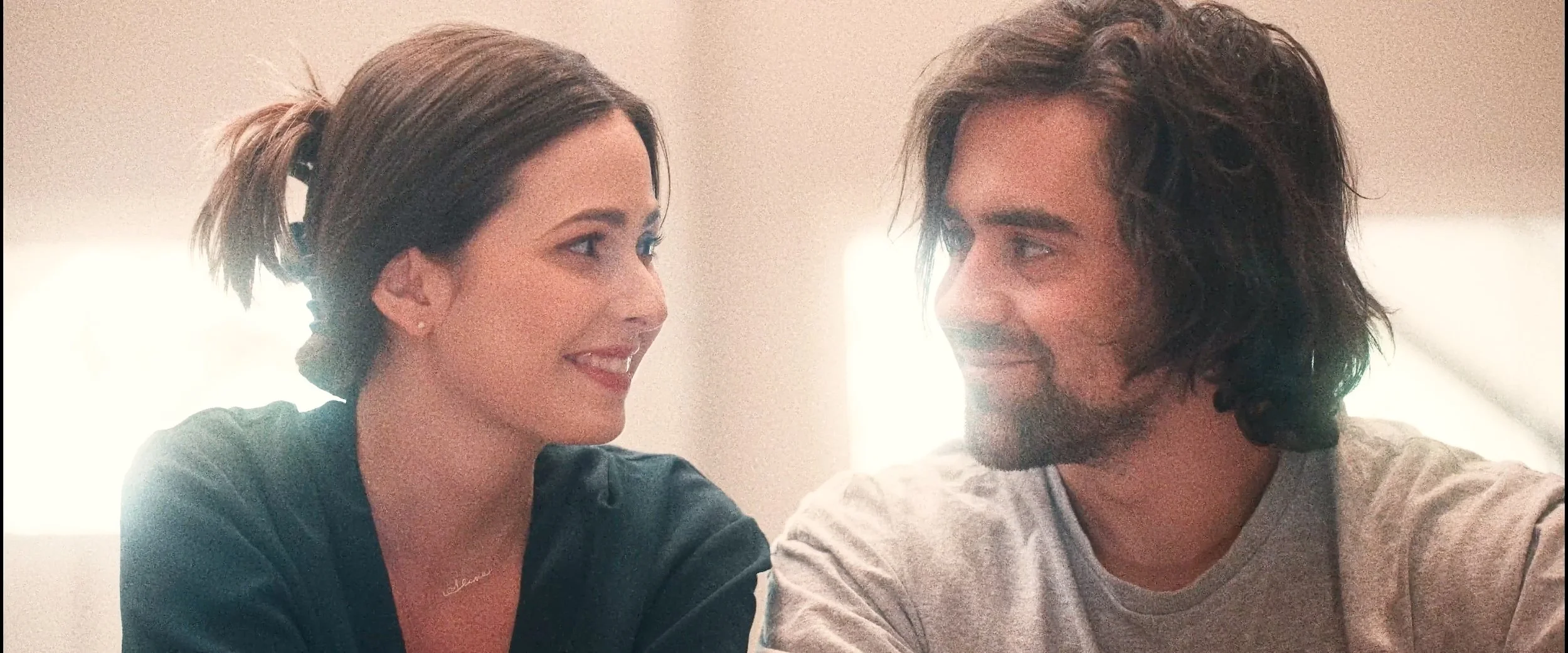 A woman and man are sitting close together, looking at each other and smiling in an indoor setting.