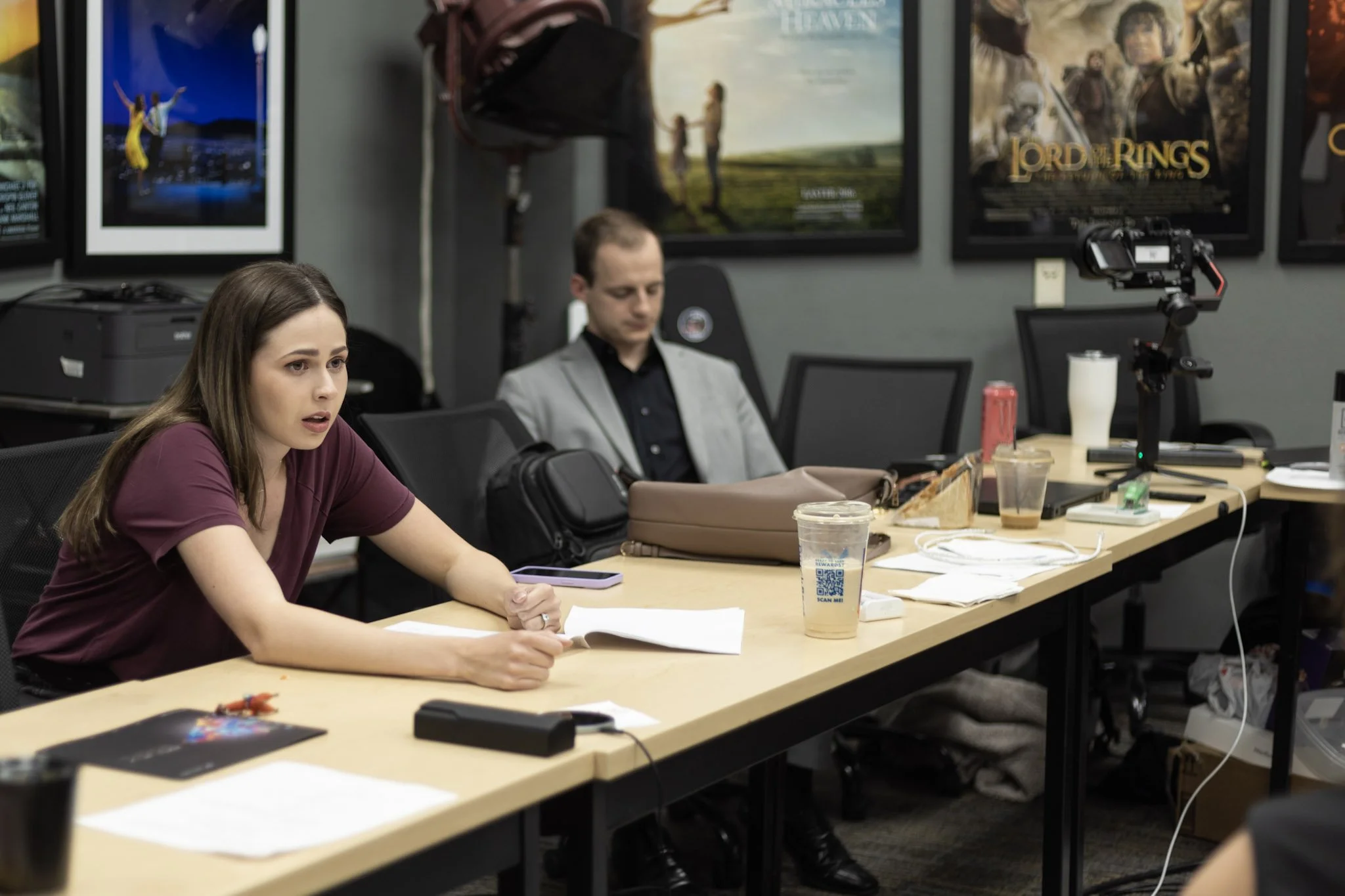 Two people sitting at a cluttered conference table, with posters and screens on the wall behind them, including one for 'Lord of the Rings'.
