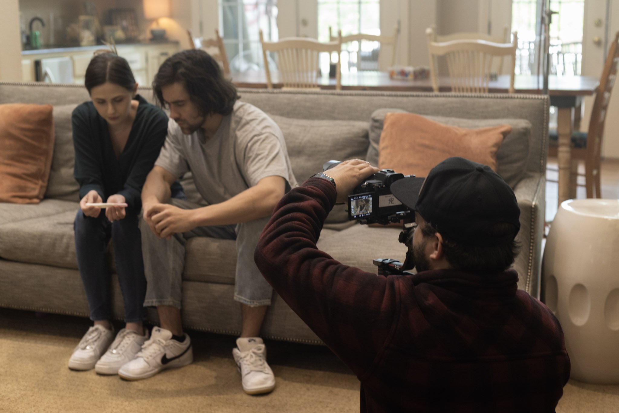 Two young adults sitting on a beige couch having a conversation while a man behind them films or photographs them with a camera.