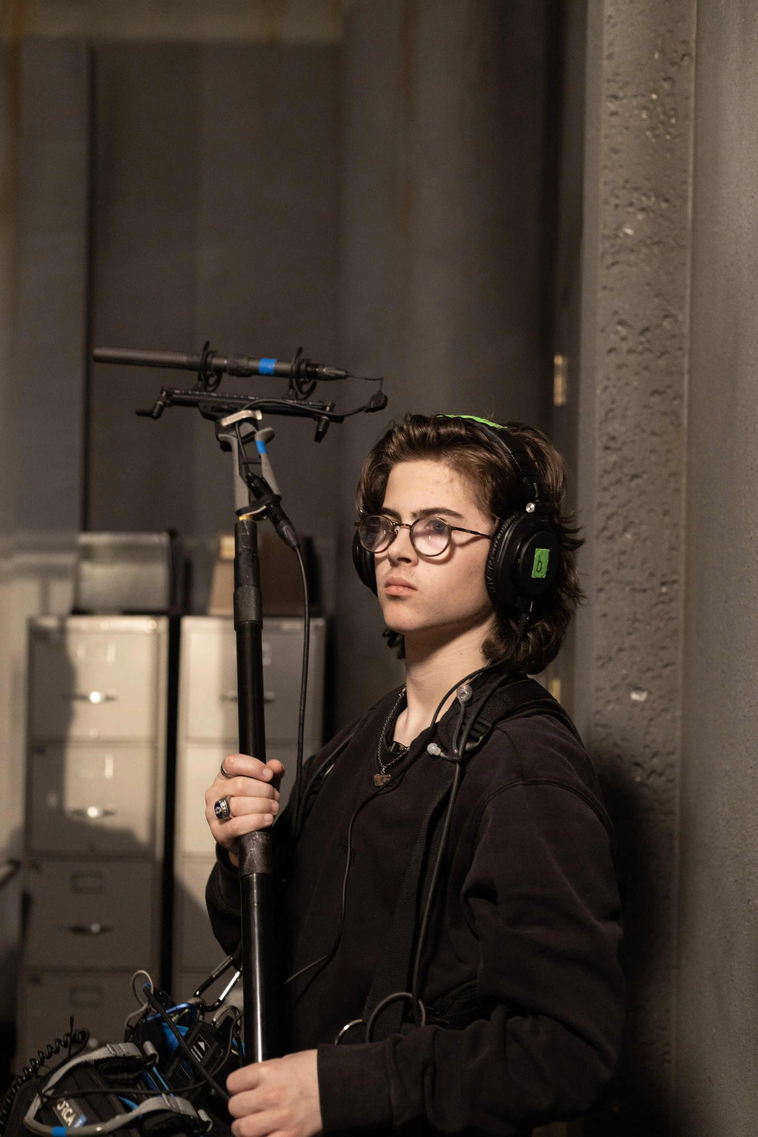 A young person with glasses and long dark hair wearing headphones and holding a boom microphone, standing against a gray wall with file cabinets in the background.