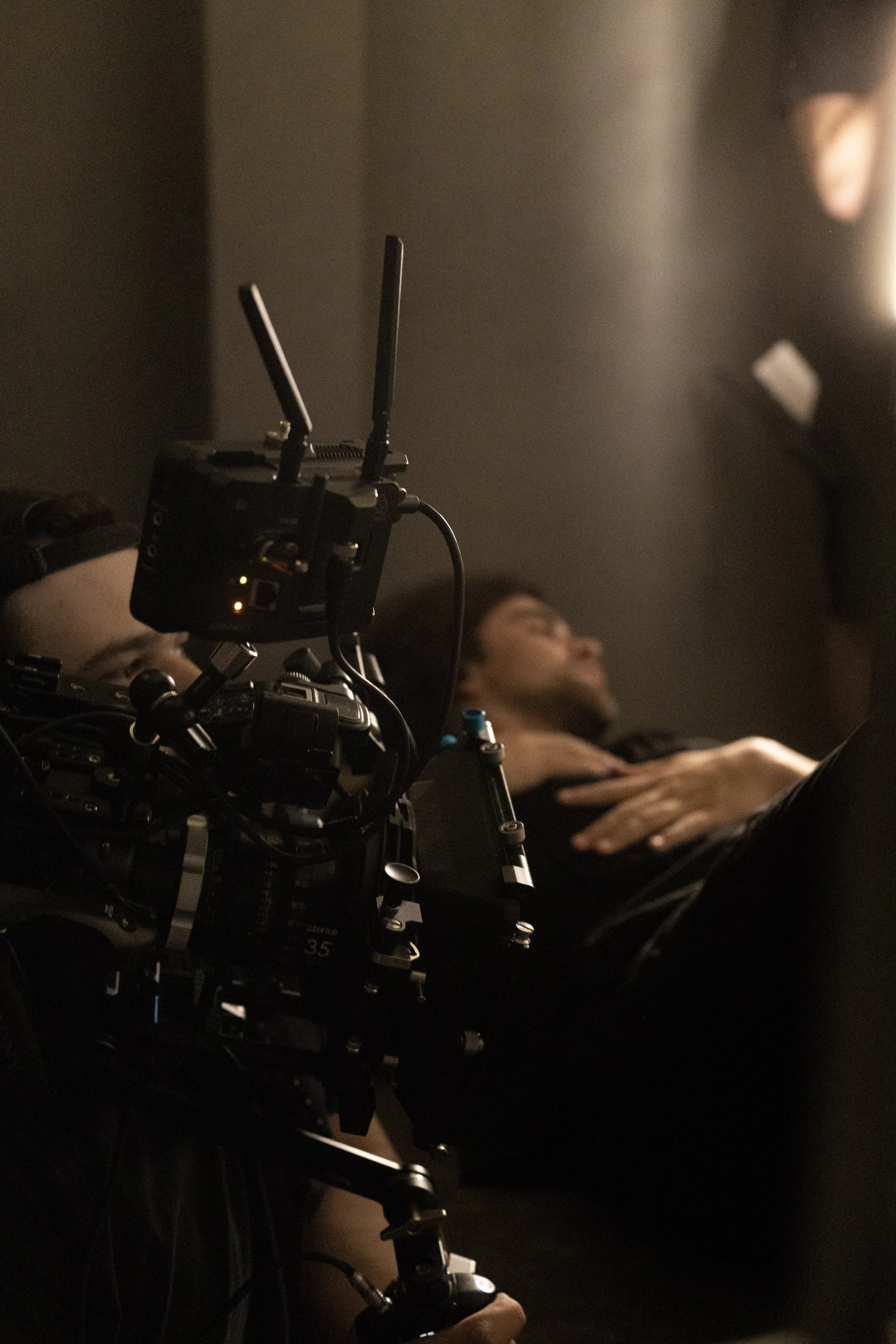 Filmmaker operating a camera while a woman is lying down with her eyes closed, possibly on a set or during a shoot.