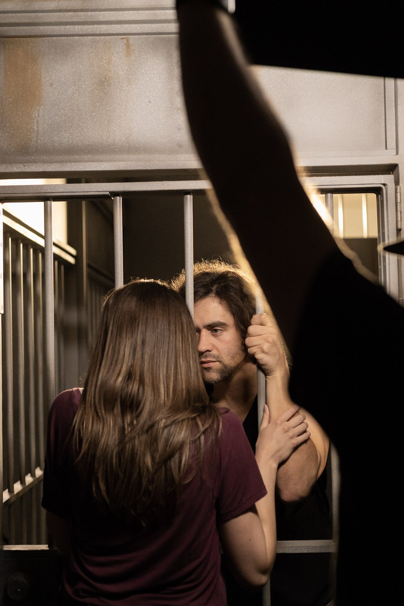A man and woman are close together behind metal bars, engaging in an intimate moment with serious expressions, inside what appears to be a jail cell.