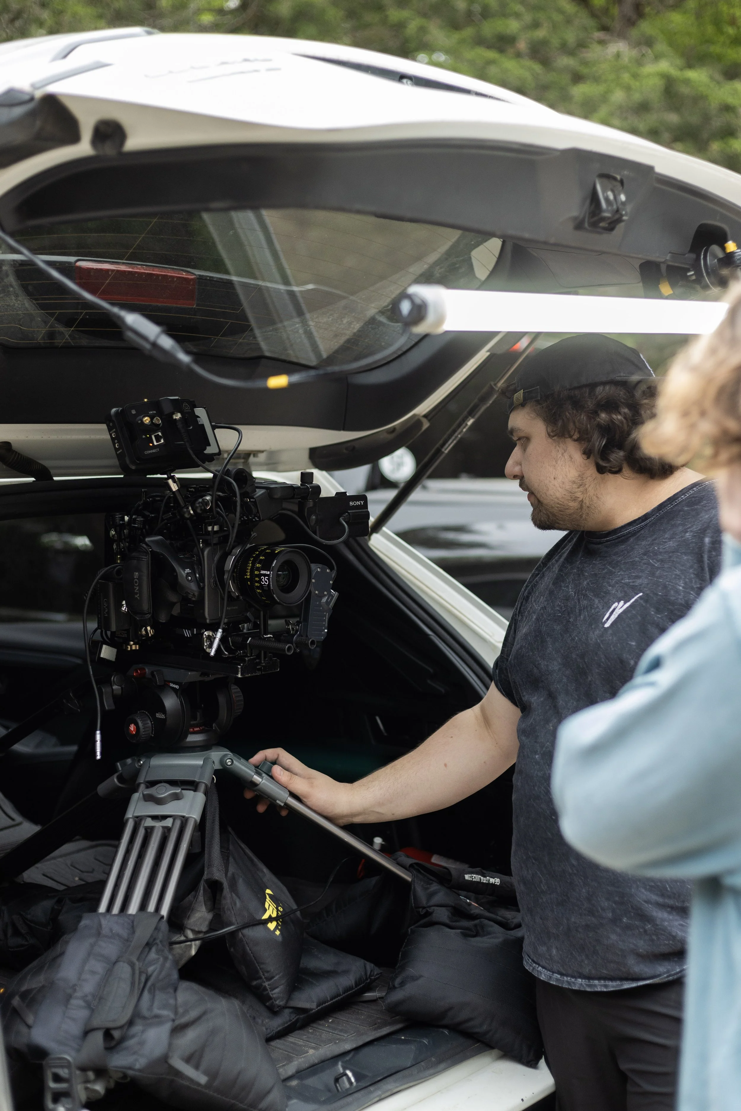 A person standing next to a camera mounted inside the trunk of a vehicle, with another person partially visible.