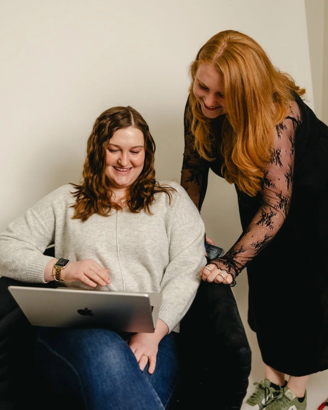 Two women smiling and looking at a laptop in a casual setting, one seated and the other leaning over.