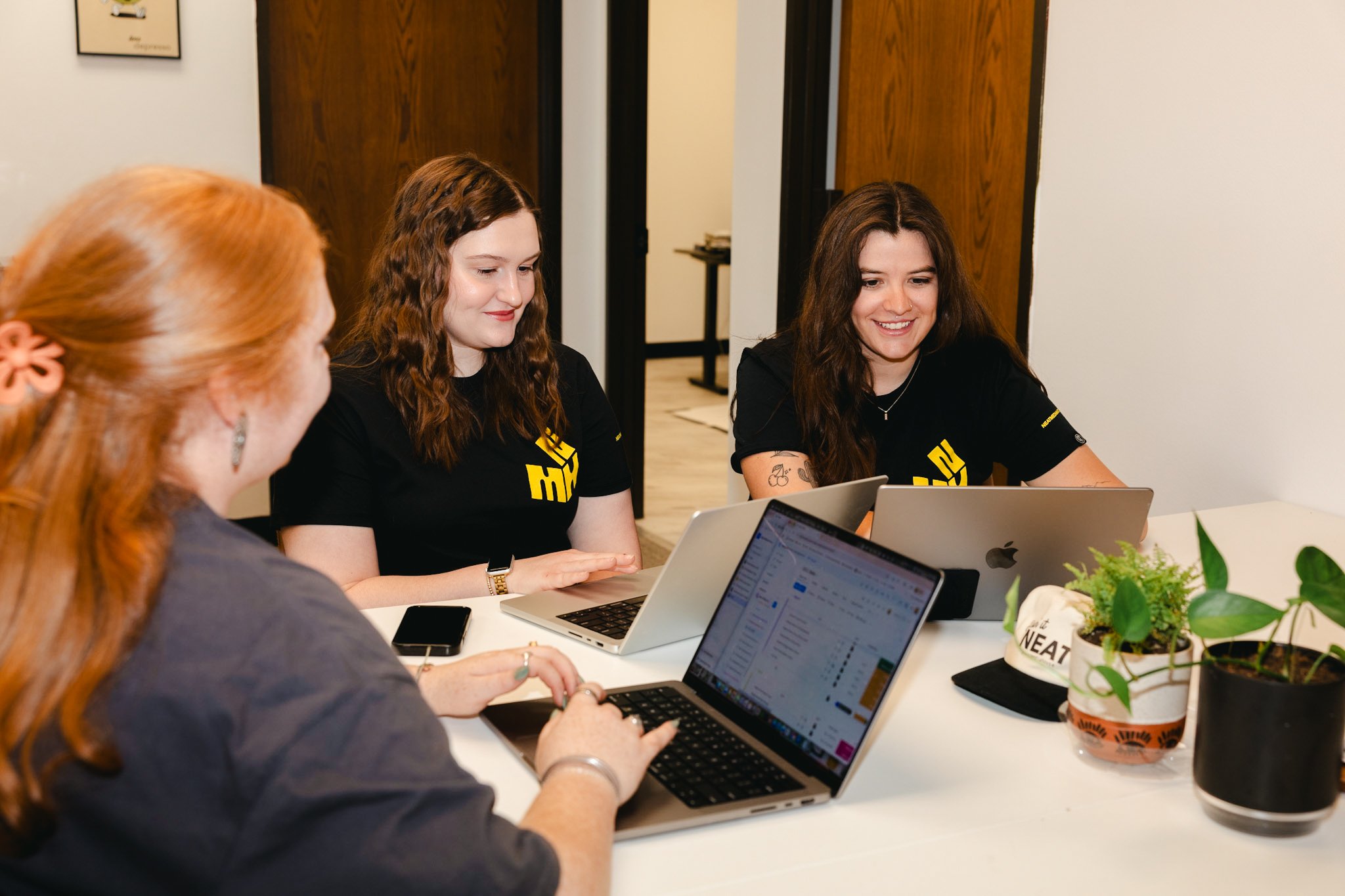Three women working on laptops at a white table in an office, with plants and a black hat nearby.