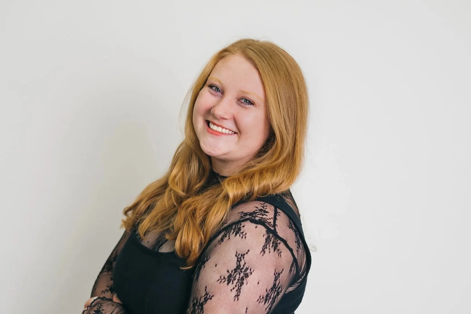 A smiling woman with curly red hair, a nose ring, and wearing a cream-colored blazer over a black top against a plain background.