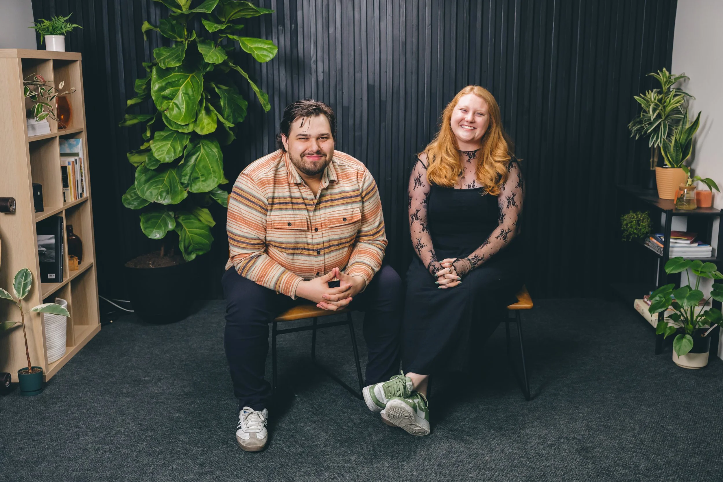 A man and woman sitting on chairs in a room with plants and bookshelves, smiling at the camera.