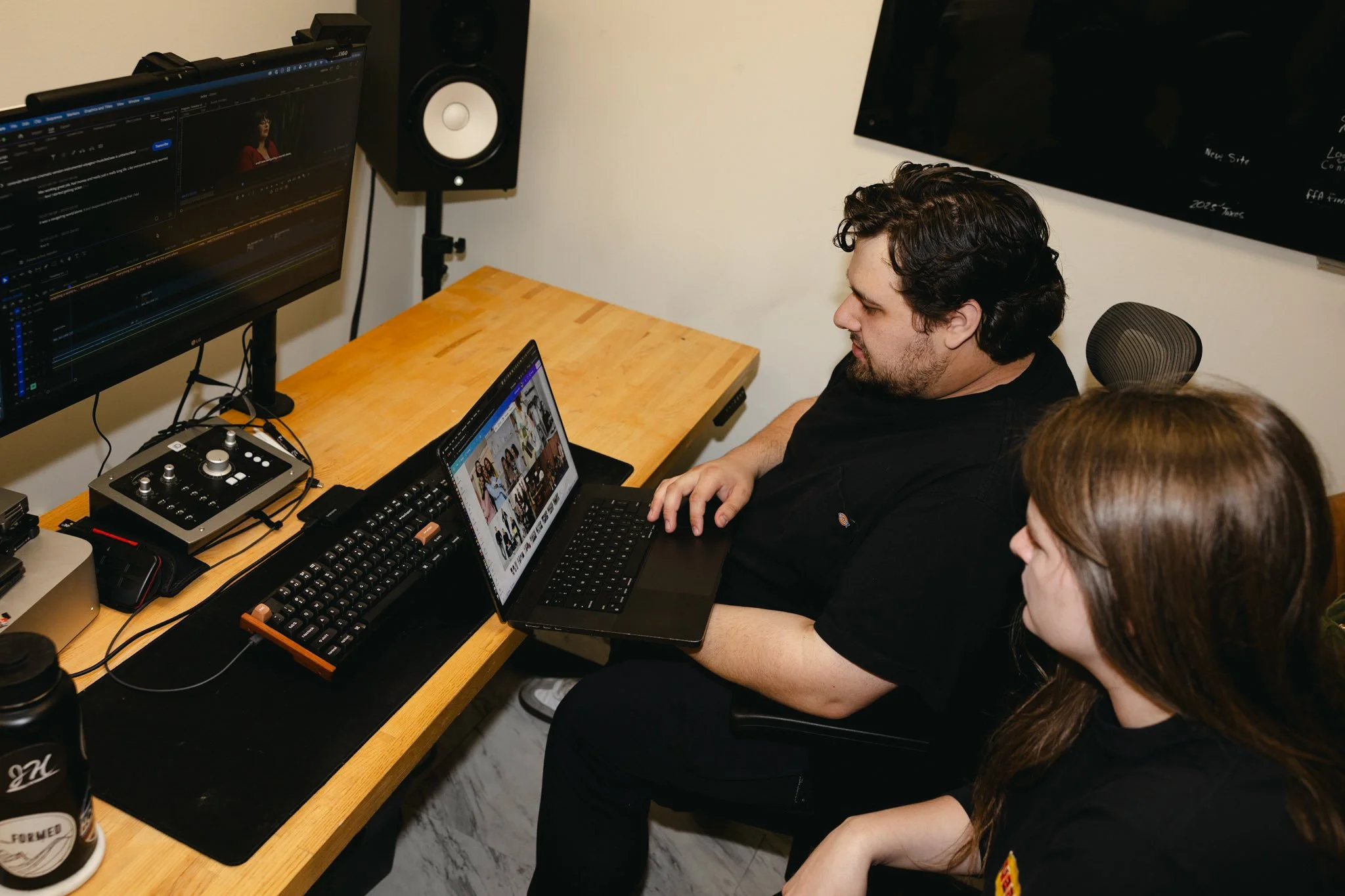 Two people working on a video editing project at a wooden desk with a computer monitor, a laptop, and audio equipment, in a room with white walls, a TV screen, and a speaker.